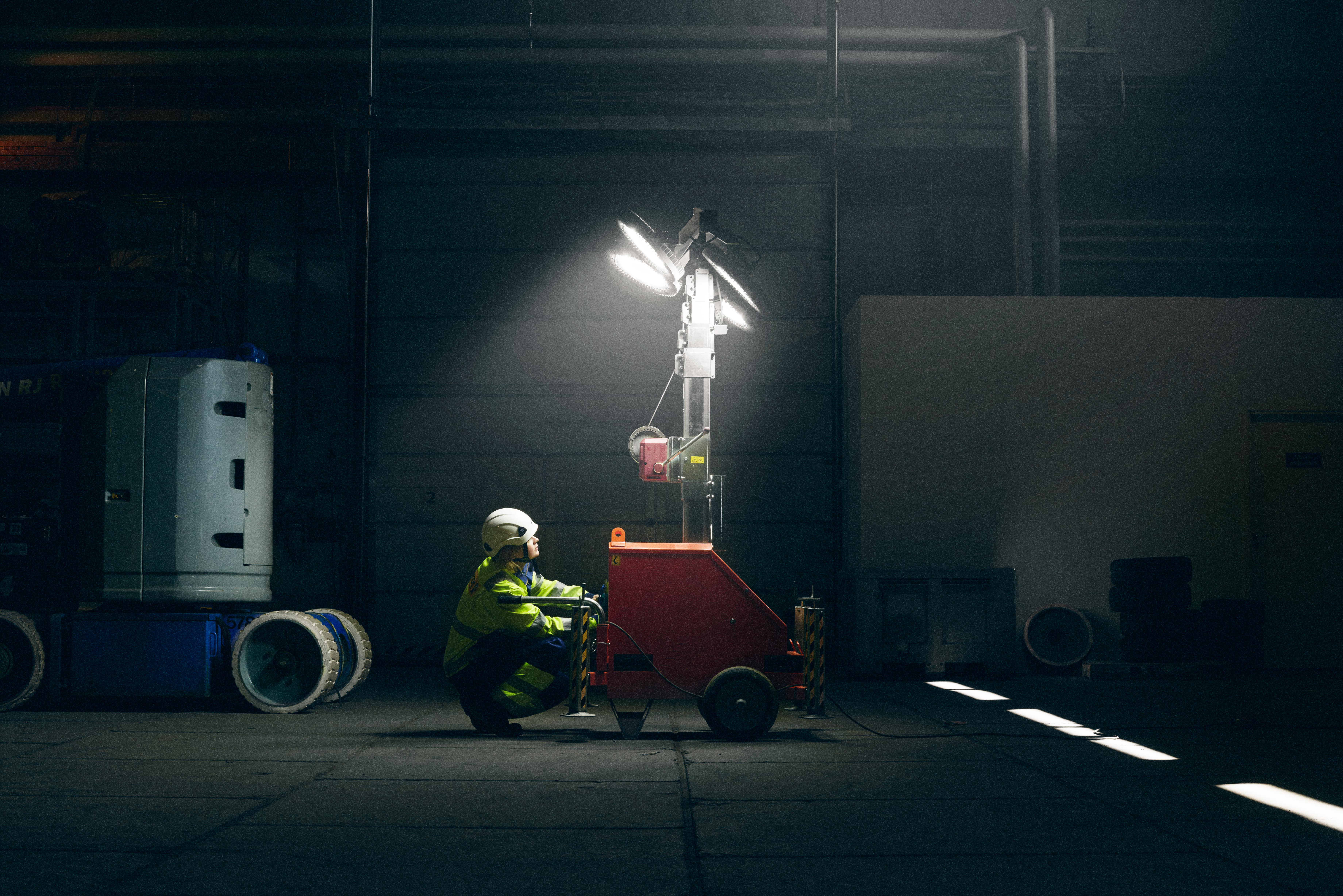 En person i varselkläder och hjälm justerar en röd mobil belysningsmast som lyser upp en mörk lagerlokal.
