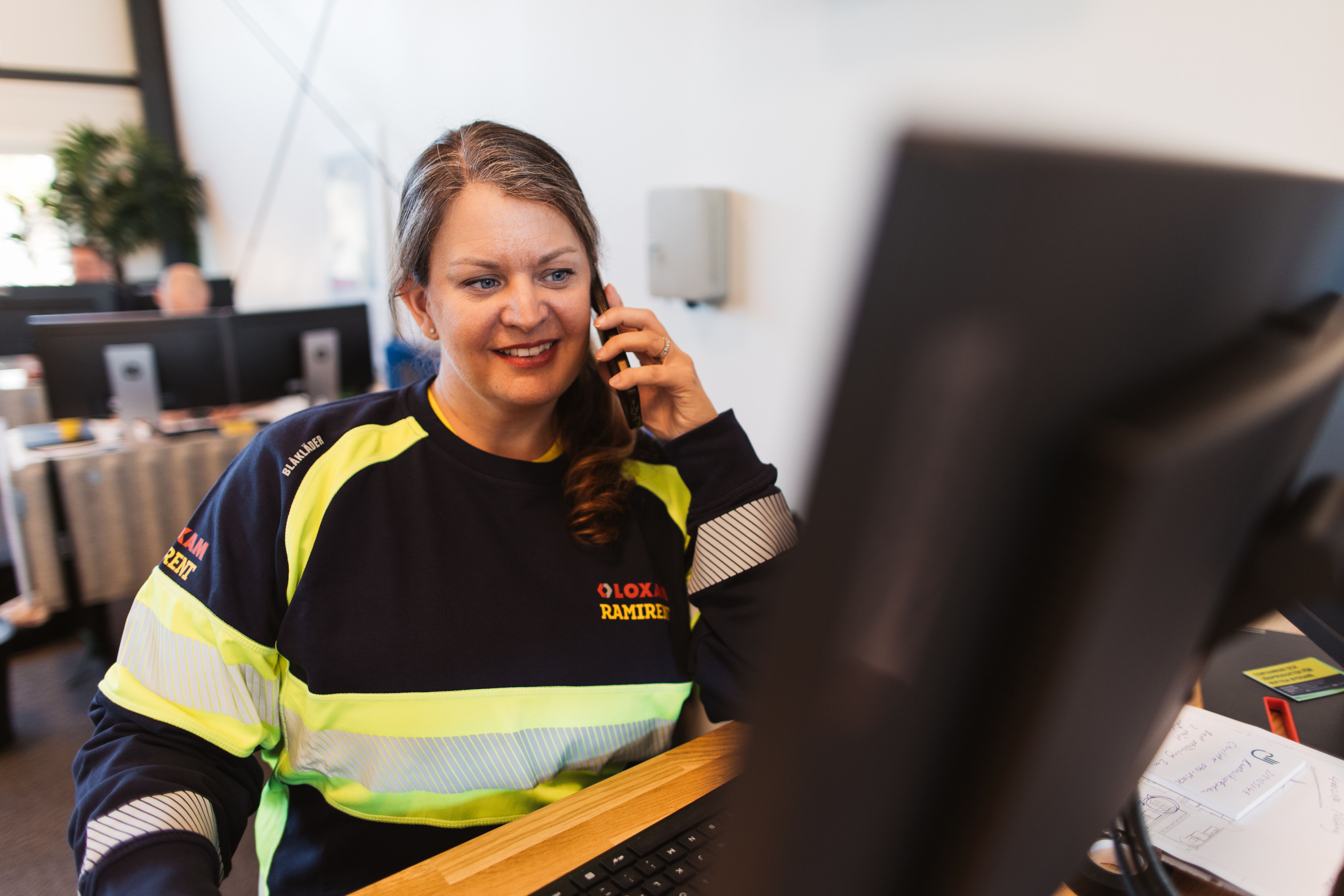 Kvinna som sitter och pratar i telefon framför sin dator, block och penna