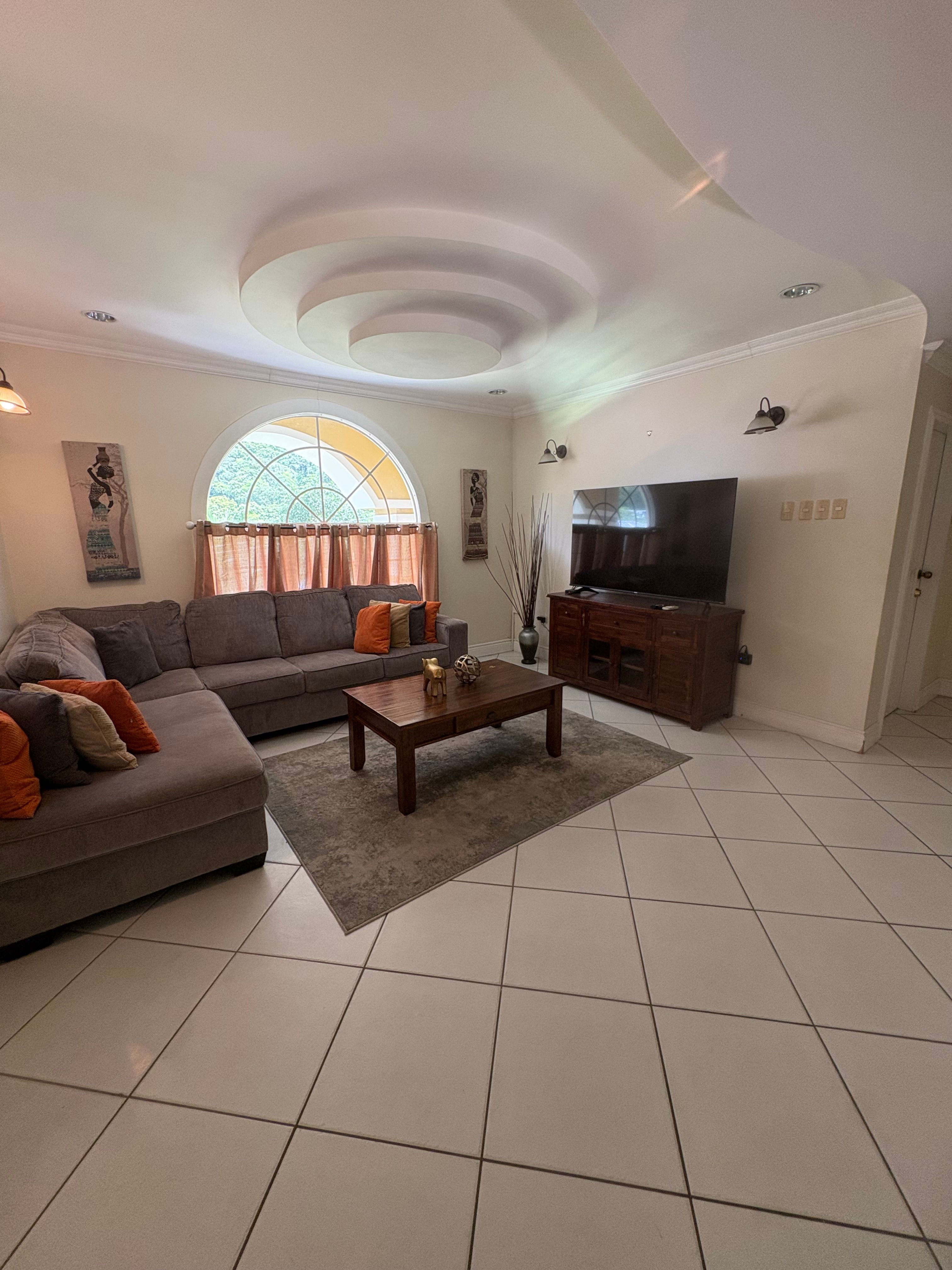 Comfortable living room with a large brown sectional sofa, coffee table, Smart TV and an arched window overlooking the greenery outside.
