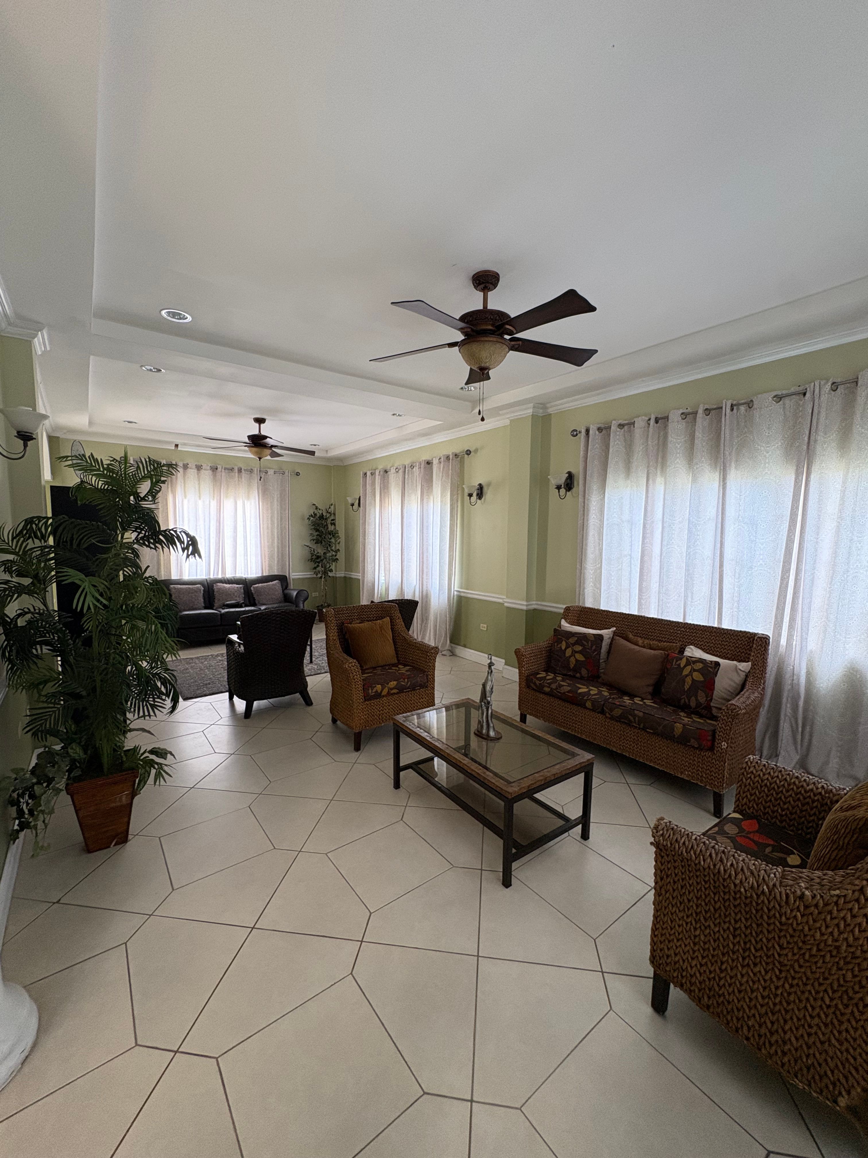 Bright and spacious sitting area with wicker sofas and armchairs, decorative plants, tiled flooring and large curtained windows.