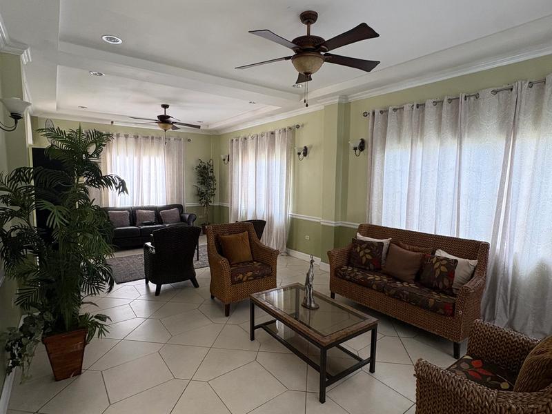 Bright and spacious sitting area with wicker sofas and armchairs, decorative plants, tiled flooring and large curtained windows.