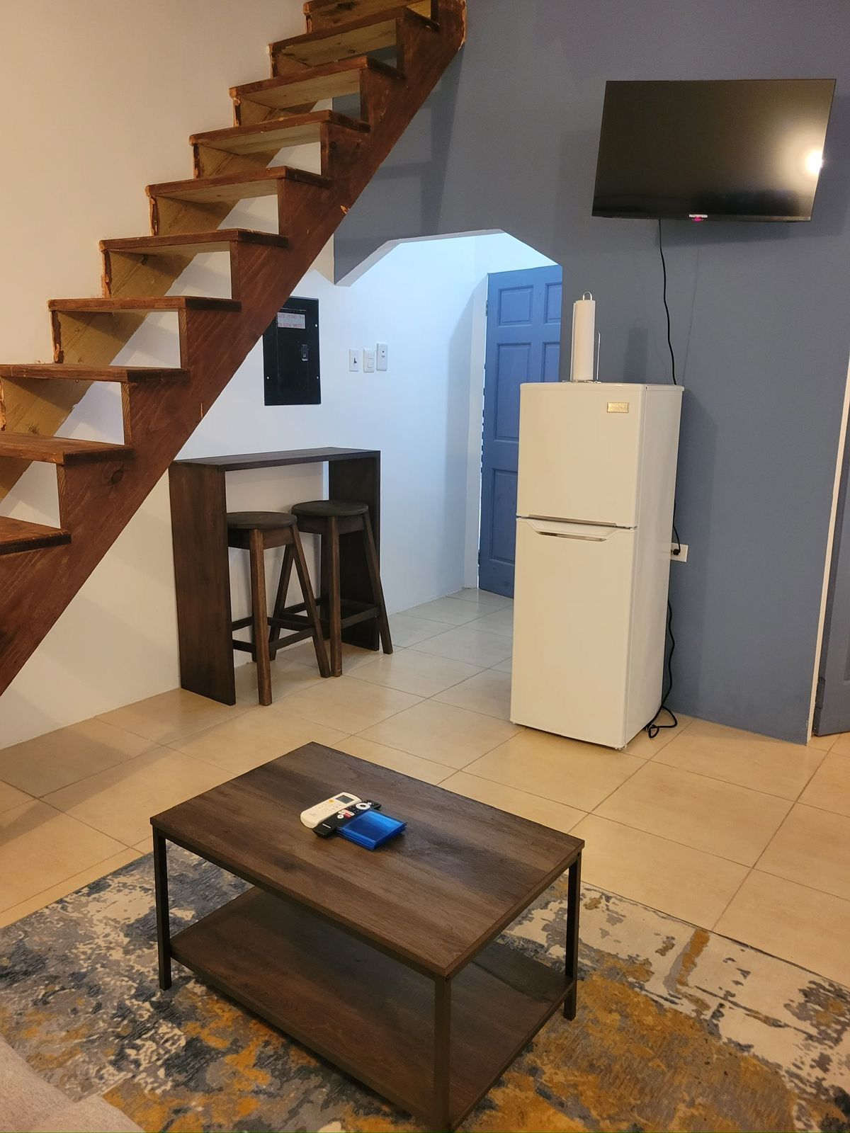 Breakfast bar with two stools beneath a wooden staircase, beside a fridge and wall-mounted television