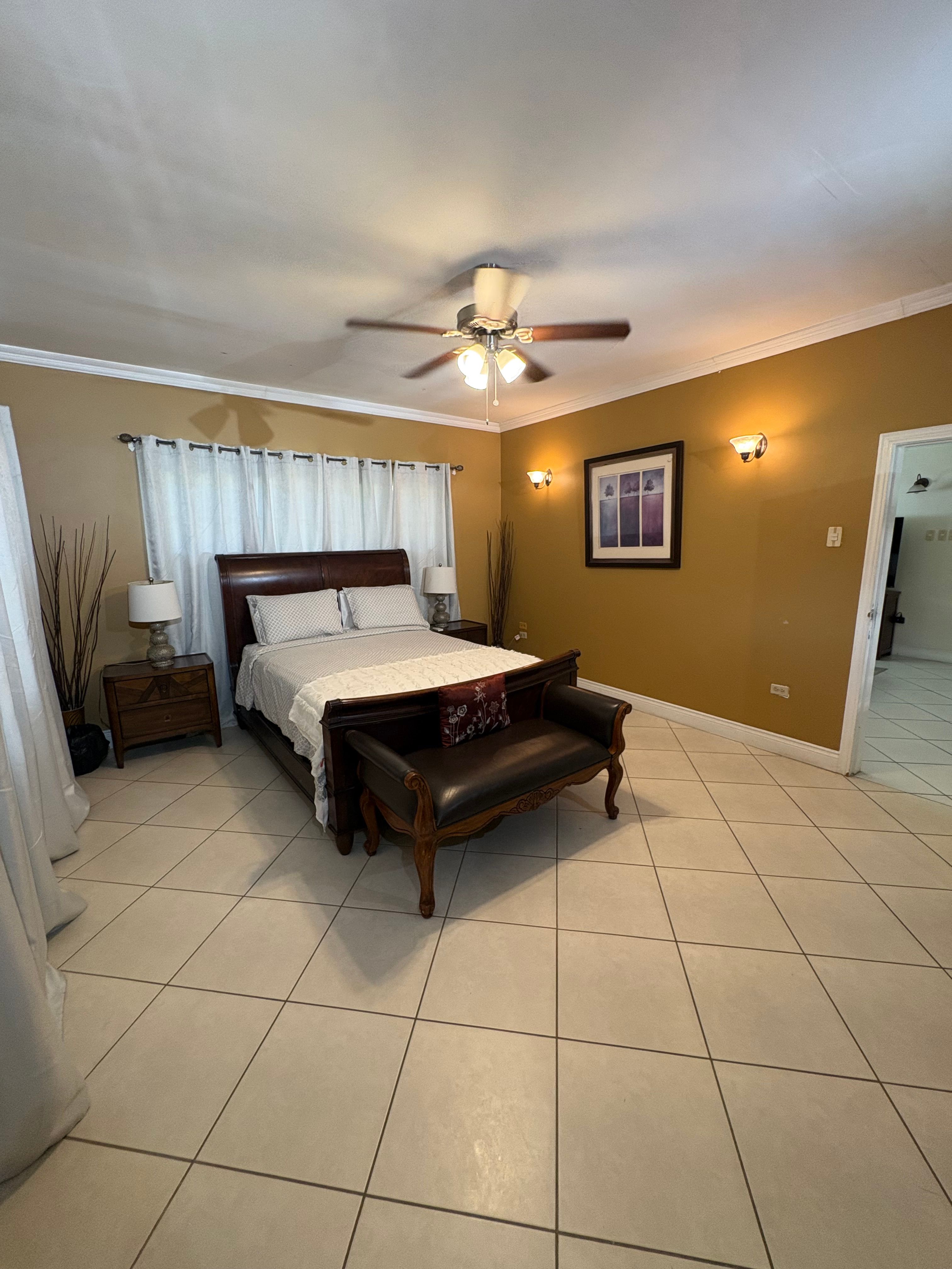 Spacious bedroom featuring a large wooden bed, matching bedside tables, warm-toned walls, tiled flooring and a ceiling fan.