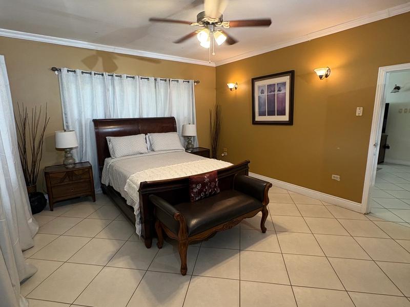 Spacious bedroom featuring a large wooden bed, matching bedside tables, warm-toned walls, tiled flooring and a ceiling fan.