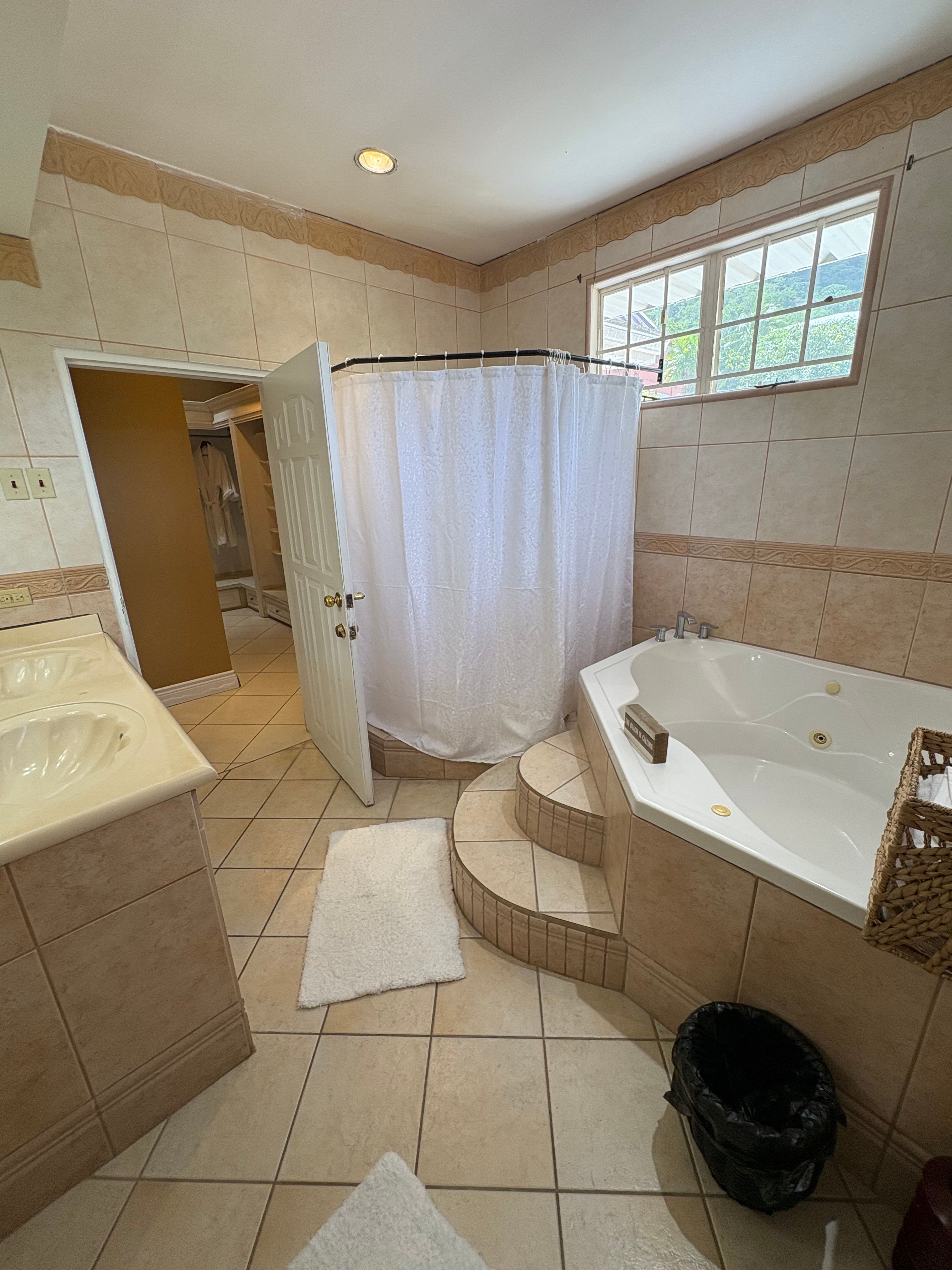 Spacious tiled bathroom featuring a corner jacuzzi bathtub, separate shower, vanity and a window overlooking the surrounding greenery.