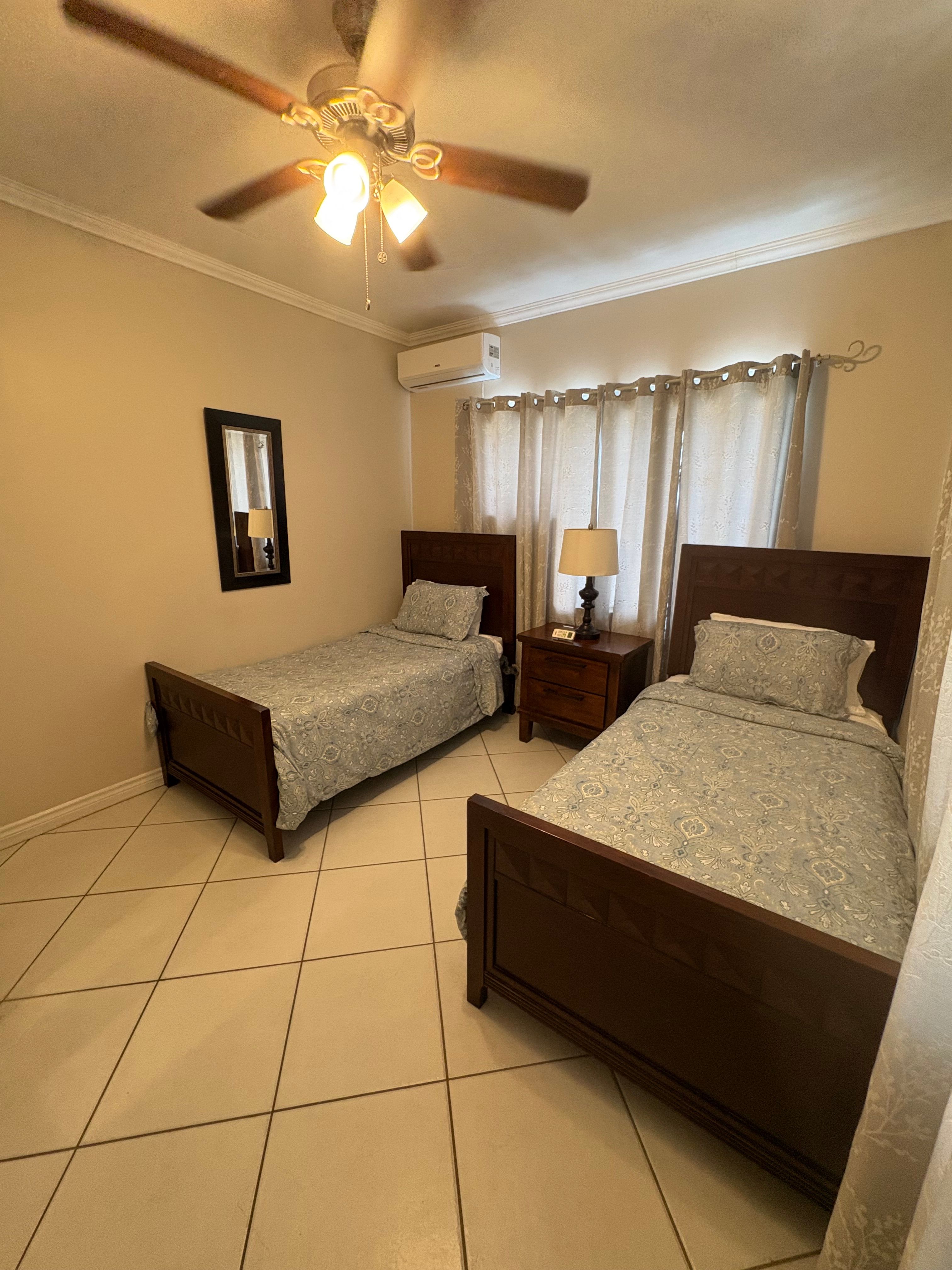 Air-conditioned guest bedroom with two single beds, wooden furniture, matching bedding and natural light through curtained windows.