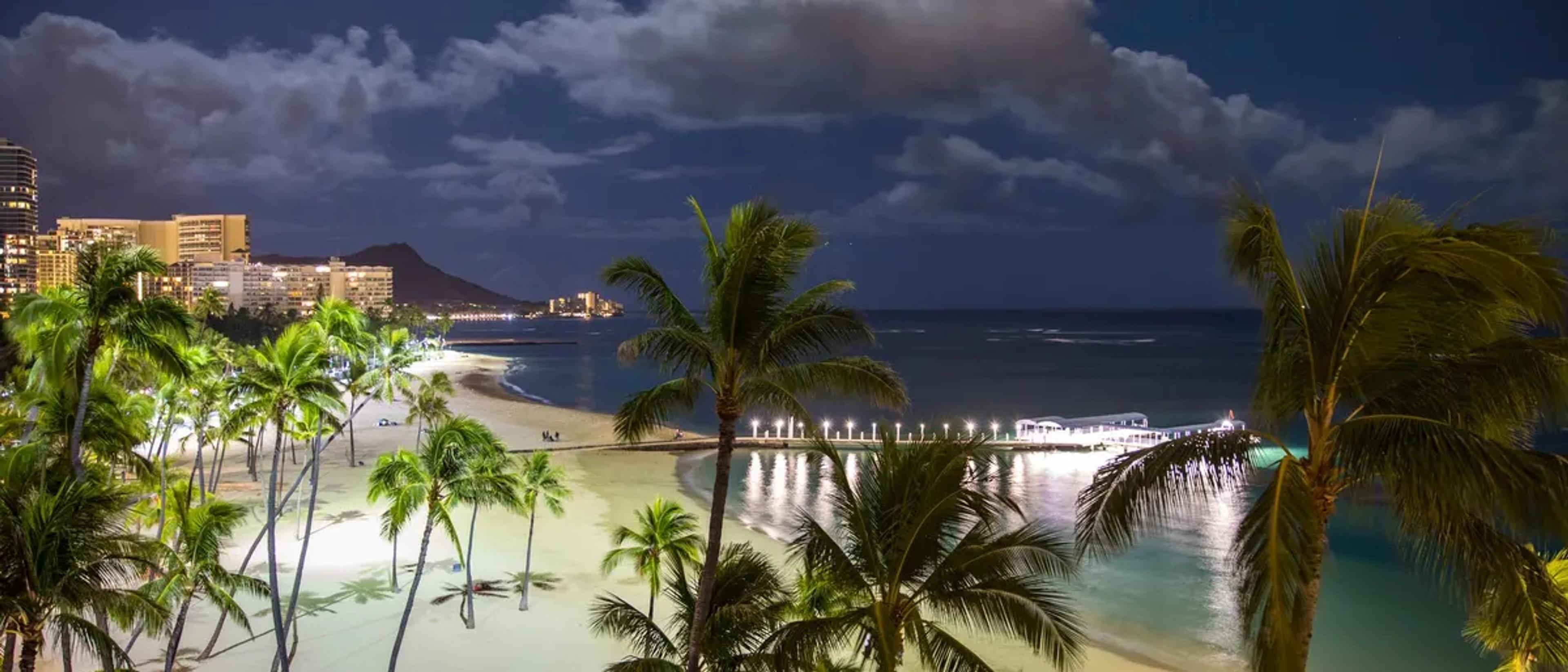 Waikiki Beach at dusk with palm trees, city lights and Diamond Head in the distance