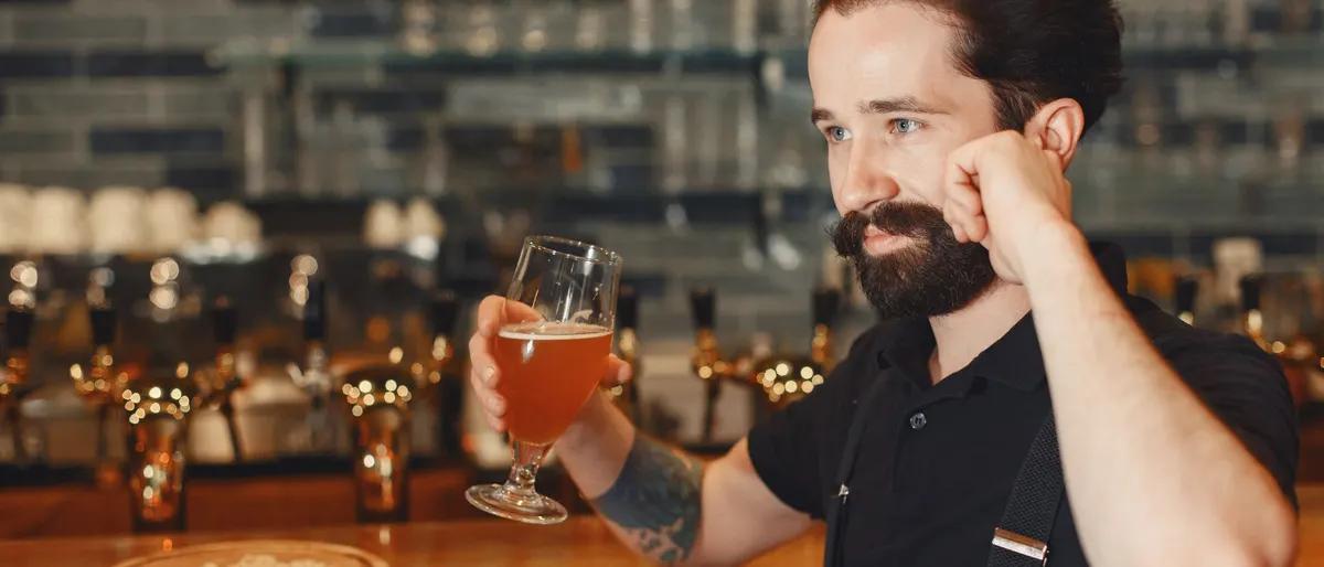 Bearded man holding a glass of beer at a bar beside a row of taps