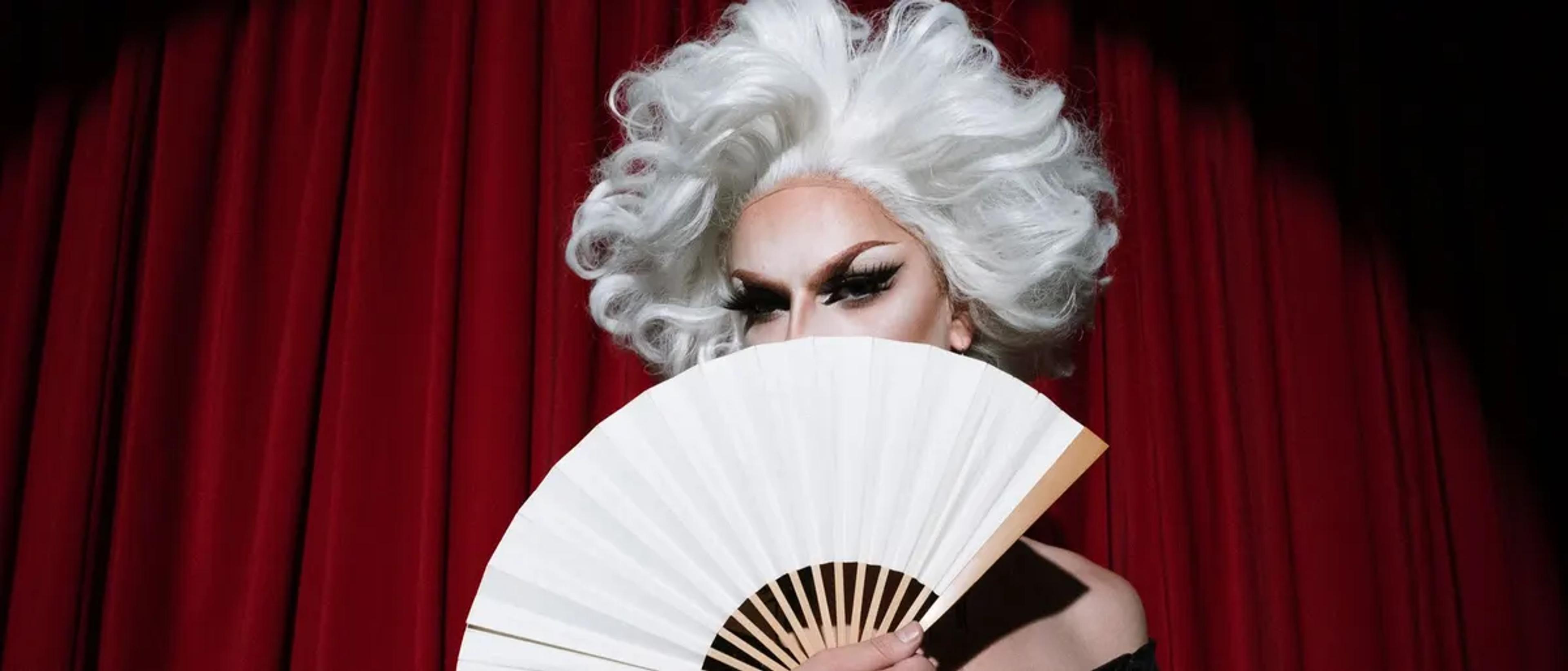 Drag performer in a white wig holding a fan in front of a red stage curtain
