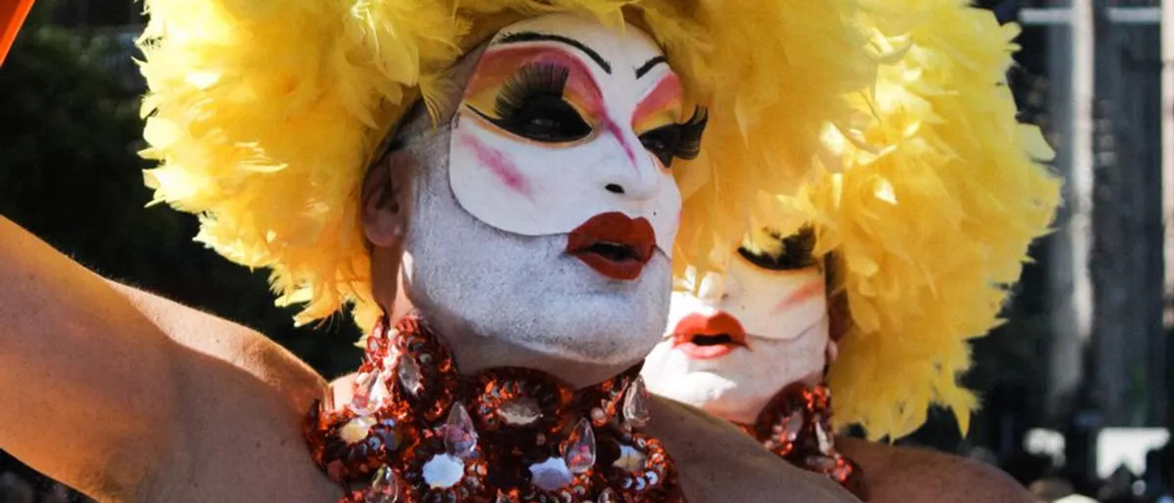 Drag queens in bold makeup and yellow feather headdresses at an outdoor Pride celebration