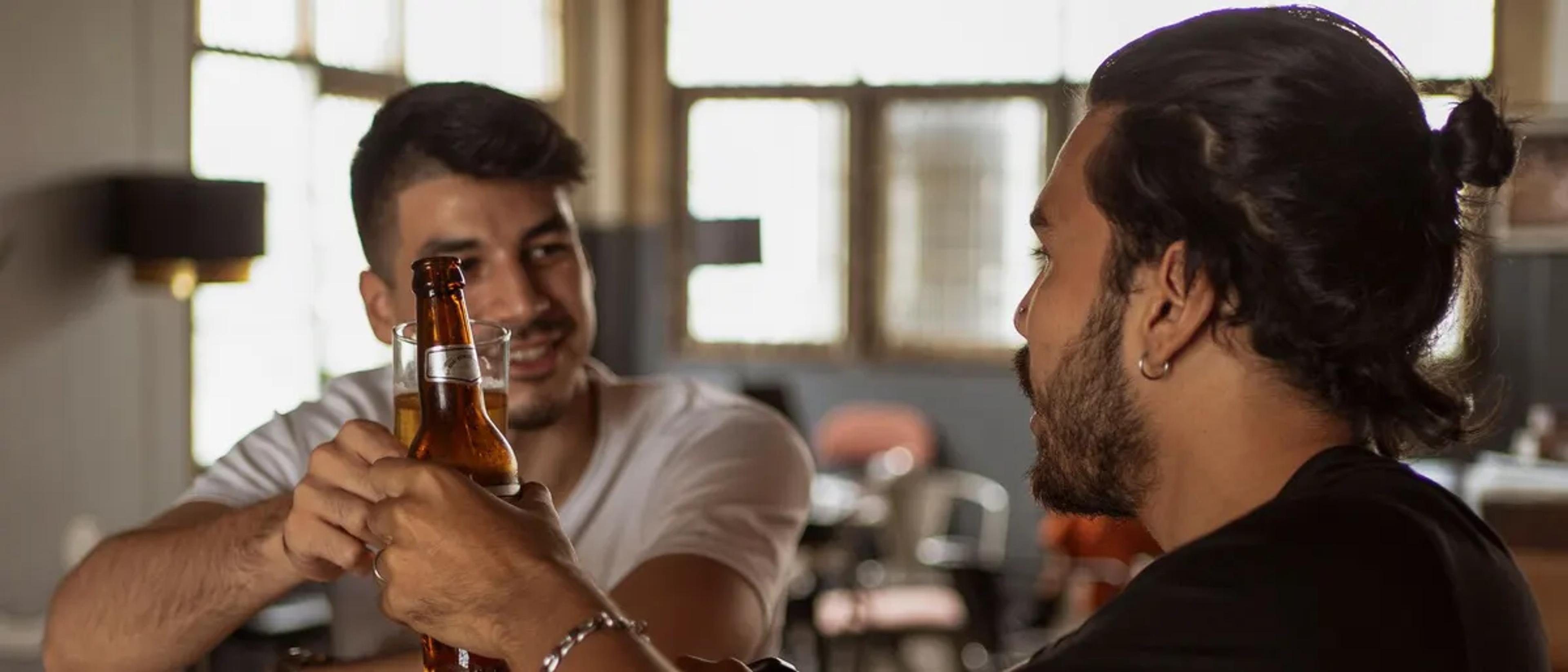 Two men clinking beer bottles at the counter of a neighborhood gay bar in the afternoon