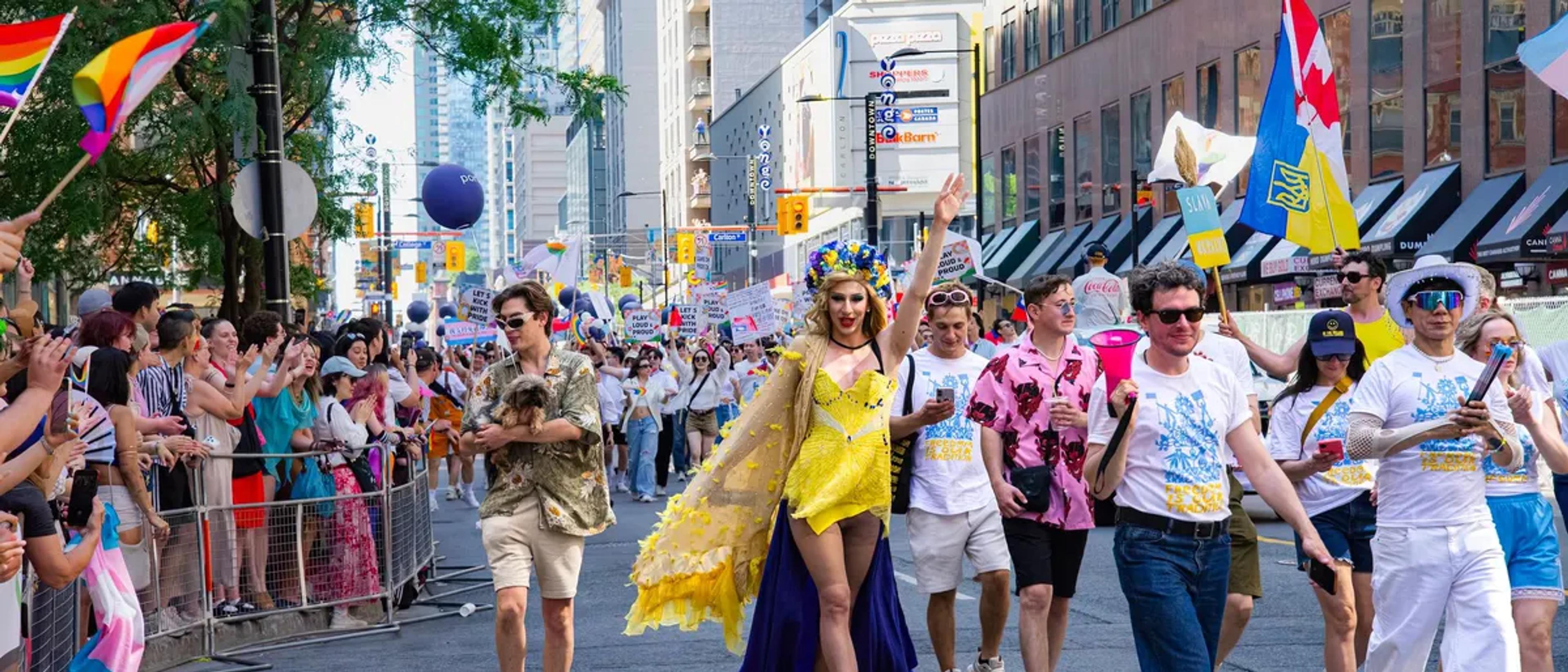 Crowds lining a downtown street waving rainbow and trans pride flags as a Pride parade passes