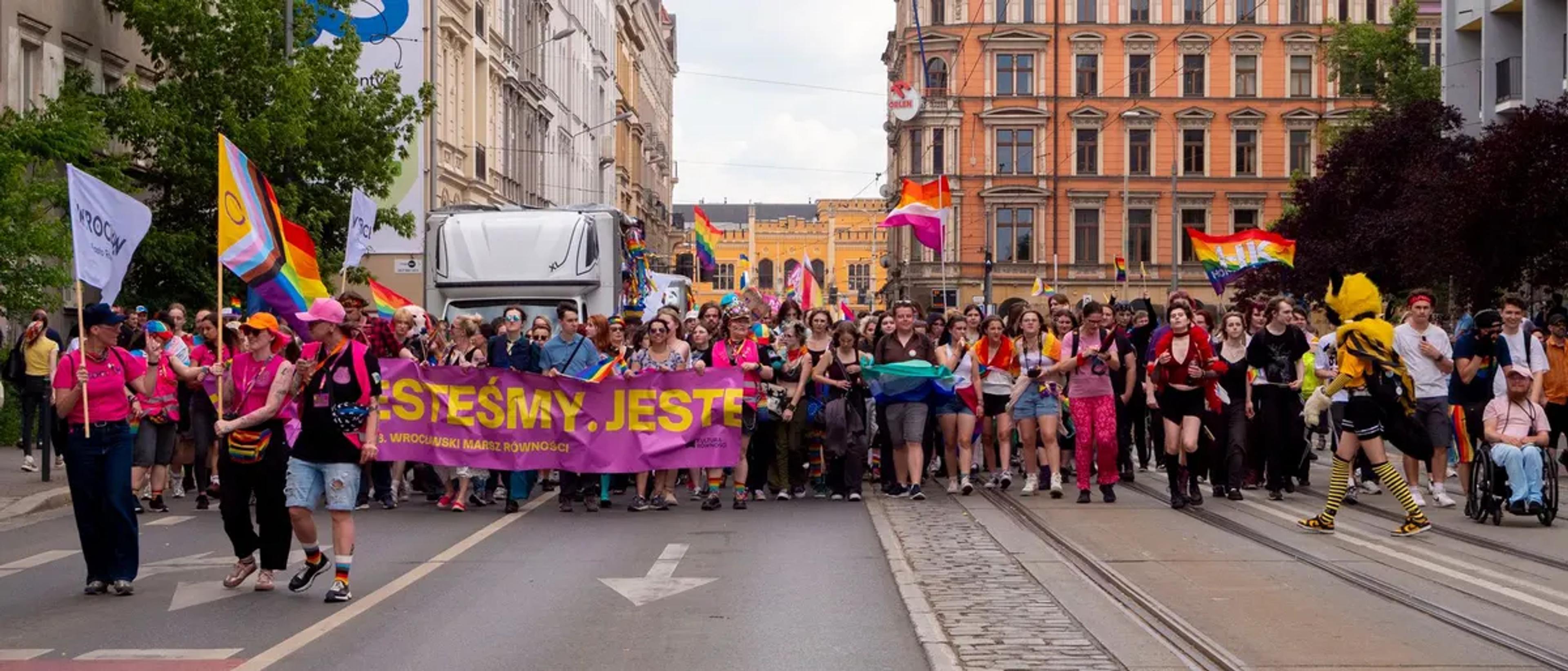 Marchers and a banner leading a Pride parade down a bright open street