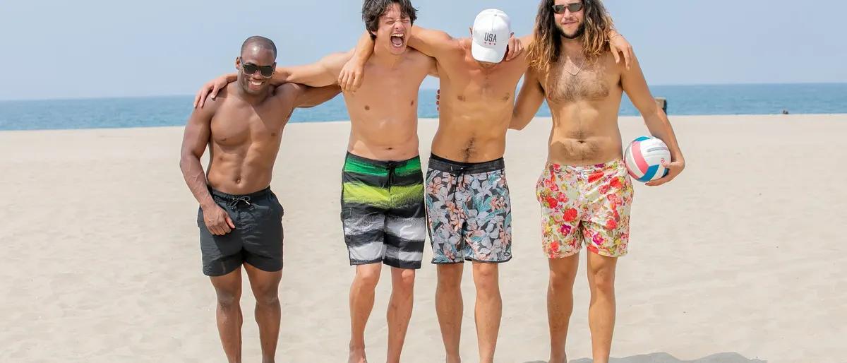 Group of men in swim trunks with arms around each other on a sunny beach holding a volleyball