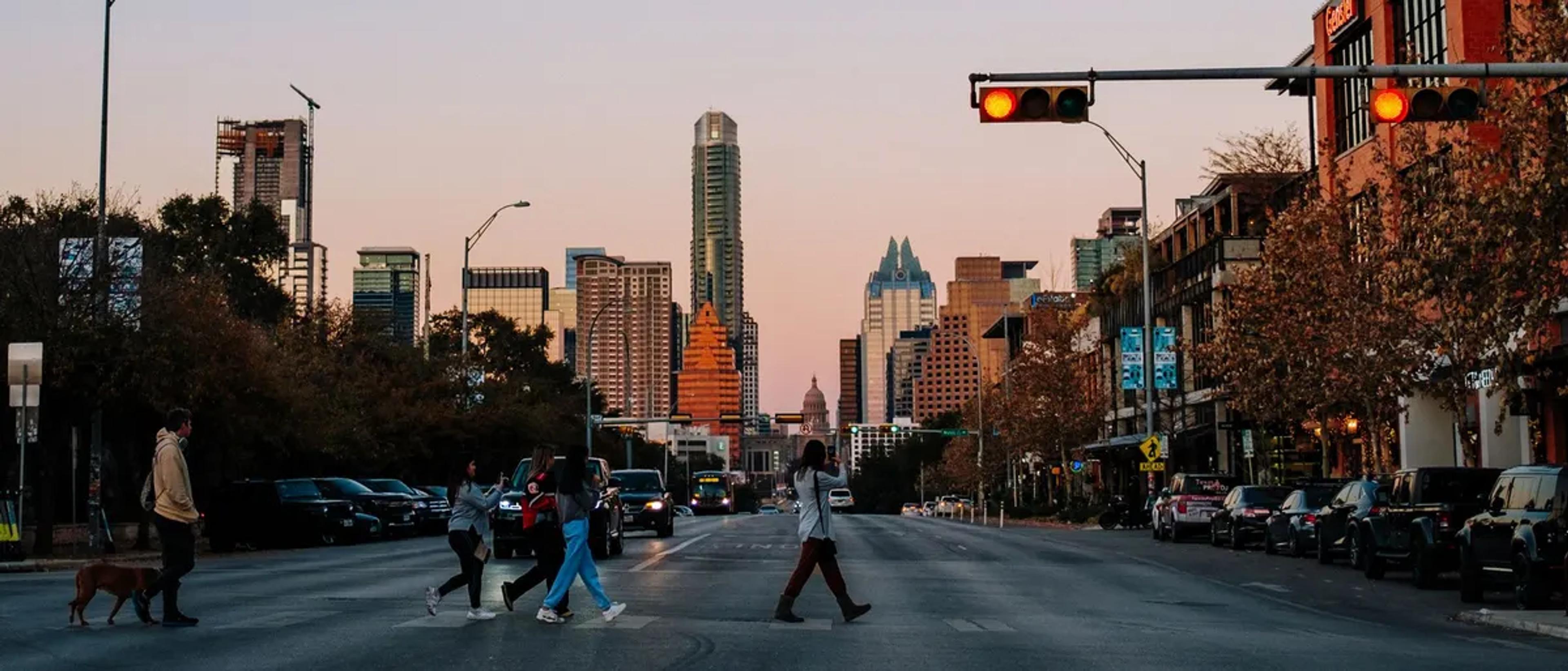 People crossing Congress Avenue in downtown Austin at sunset, with the skyline behind them