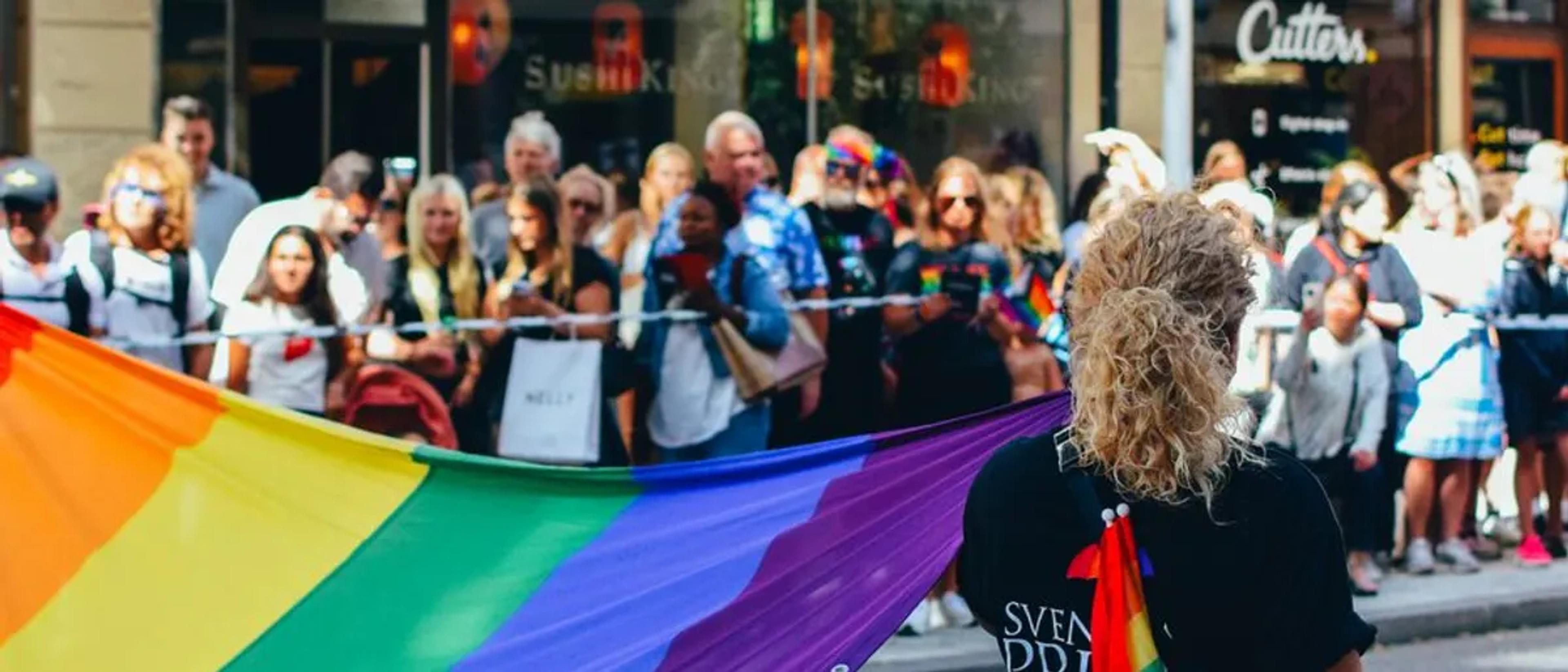 A rainbow flag carried past a lively crowd lining a downtown Pride parade route