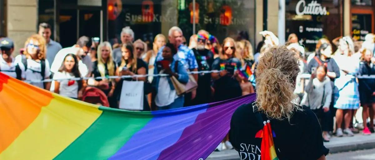 A rainbow flag carried past a lively crowd lining a downtown Pride parade route