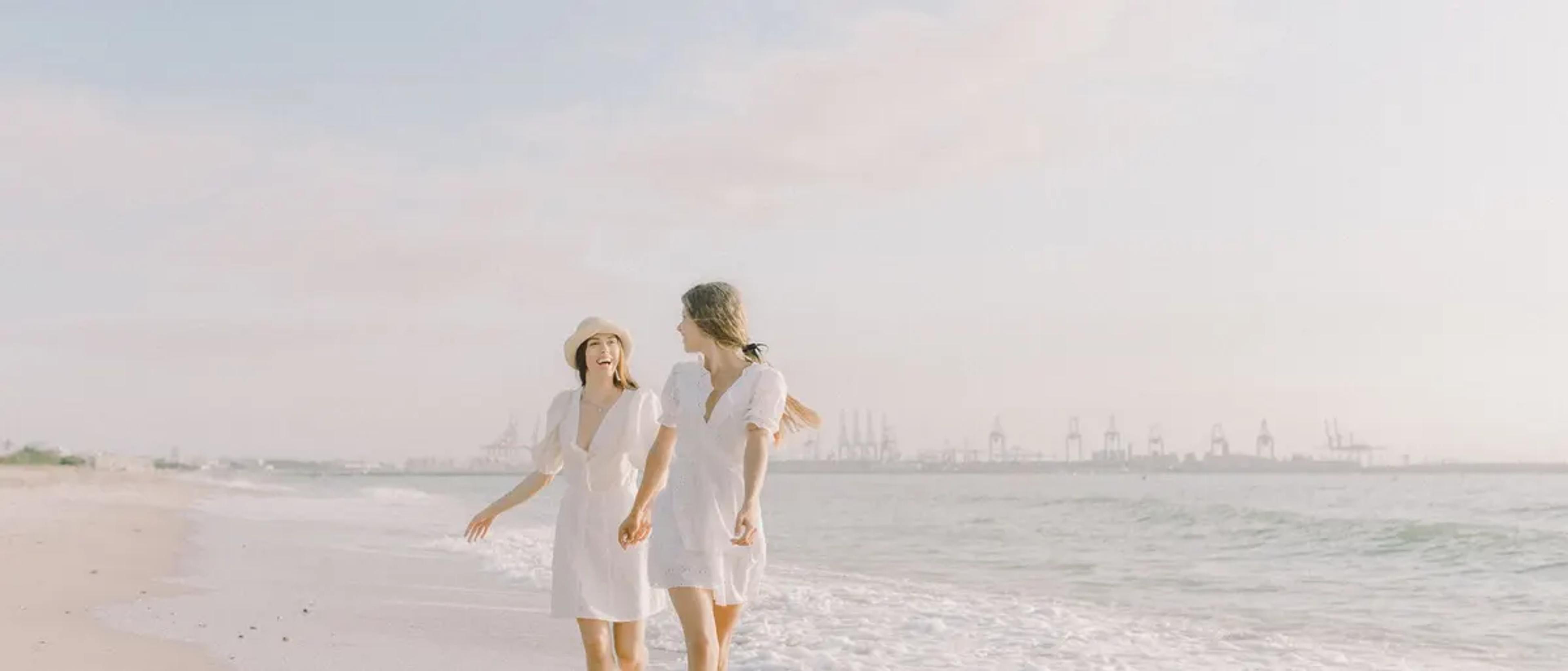 Two women in white dresses walking on a beach by the sea
