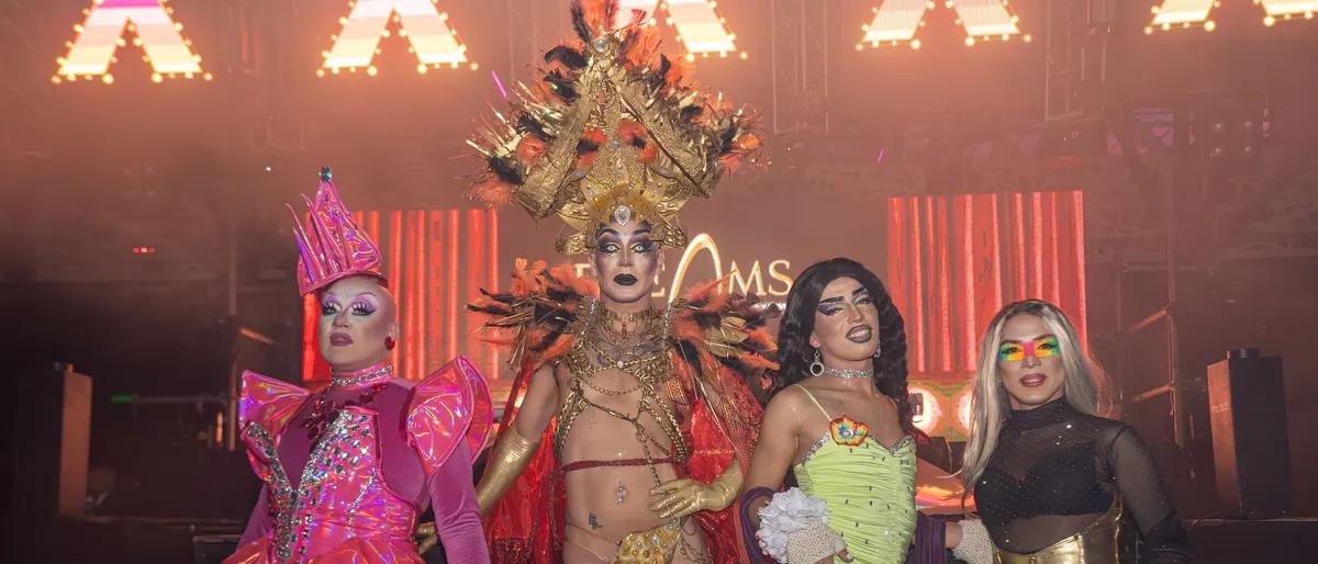 Four drag performers in bright costumes on a bar stage under pink and red lights