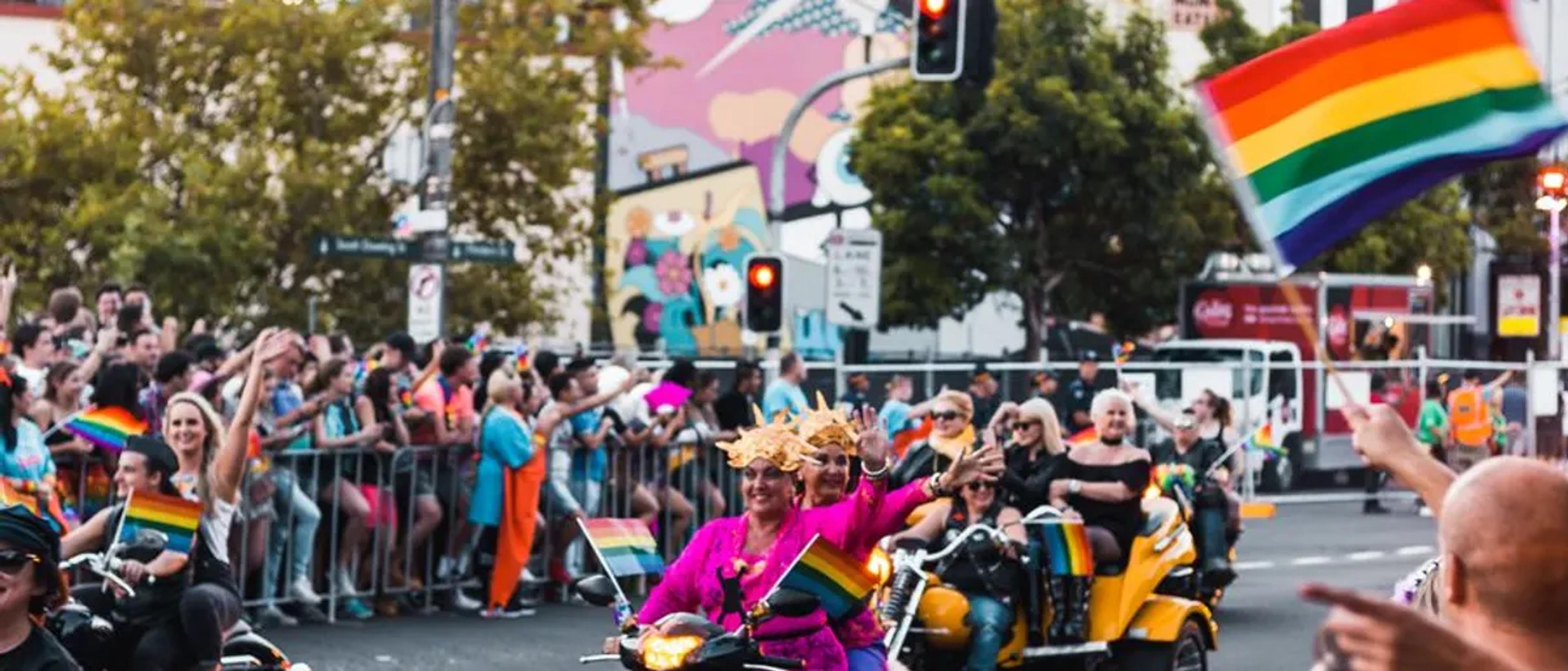 Crowds cheering behind barriers as rainbow flags fly along a sunny Pride parade route