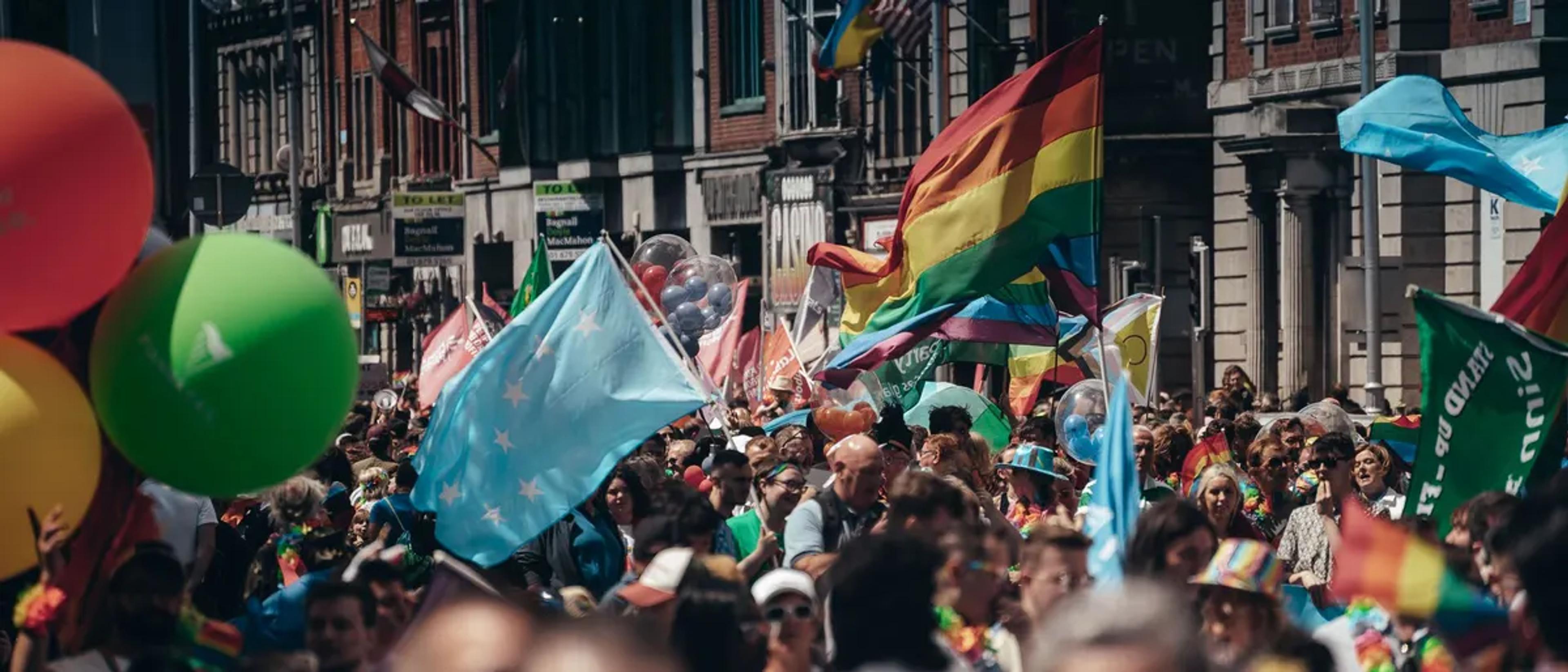 A festive Pride crowd with balloons and rainbow flags in a city square