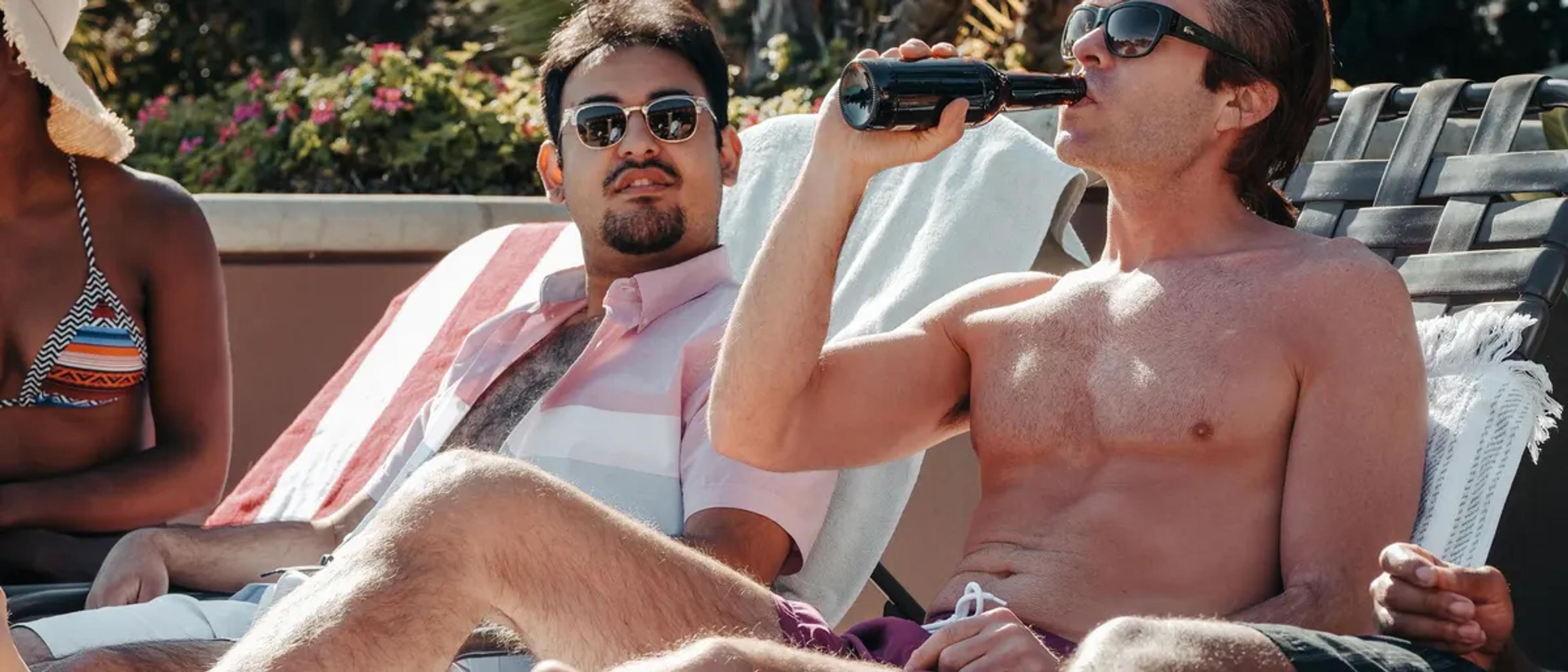 Two shirtless men relaxing on poolside loungers in the sun with a drink