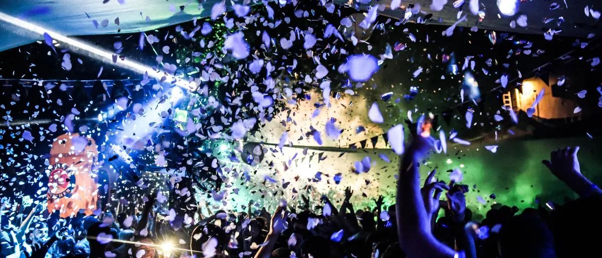 A packed dance floor under blue and green lights with confetti falling over the crowd at a circuit party