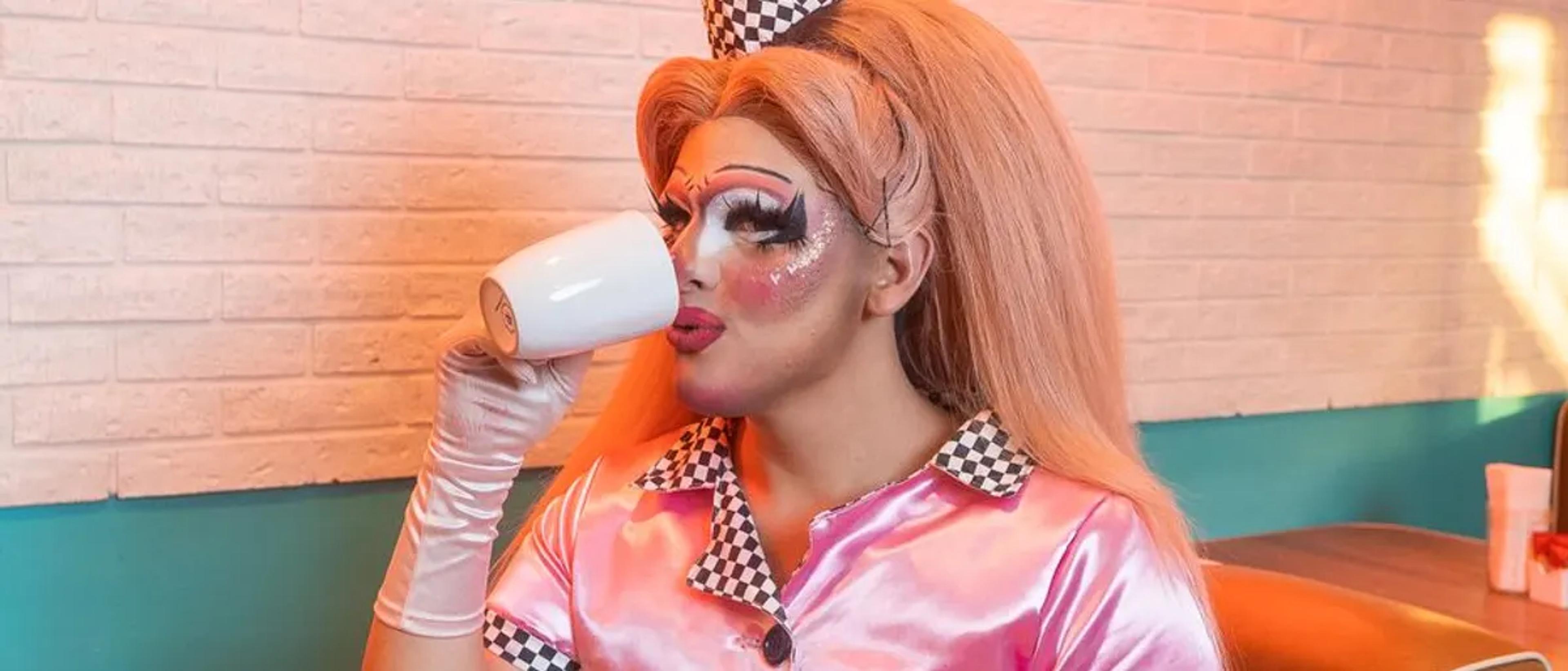 Playful drag performer sipping from a mug in a pink diner uniform at brunch