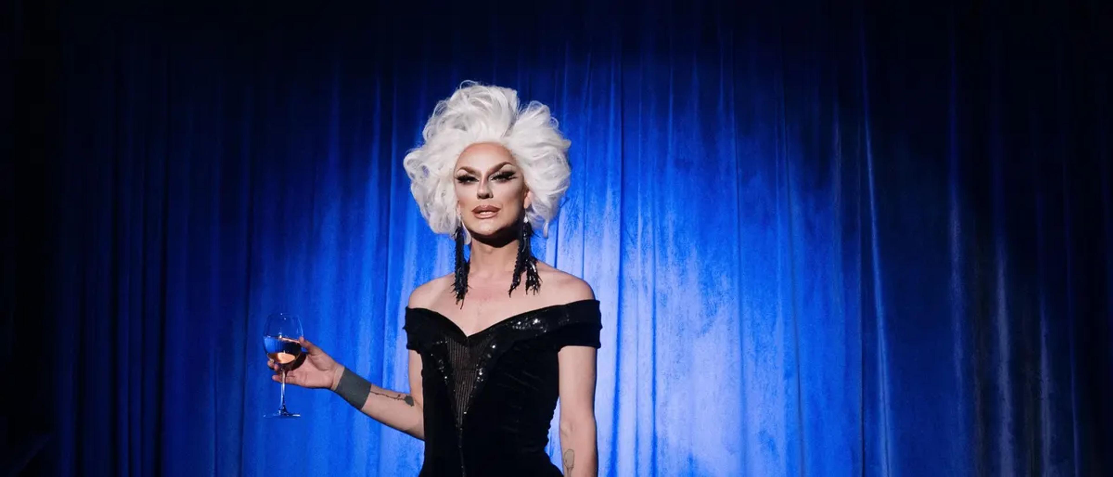 Drag performer in a white wig and black gown holding a glass of wine in front of a blue stage curtain