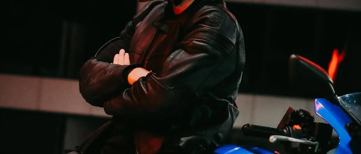 Man in a black leather jacket standing arms crossed beside a motorcycle under red light