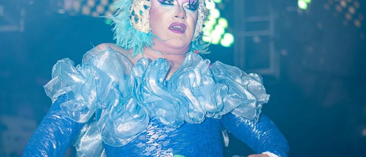 Drag queen in a blue ruffled costume performing under stage lights at a nightclub