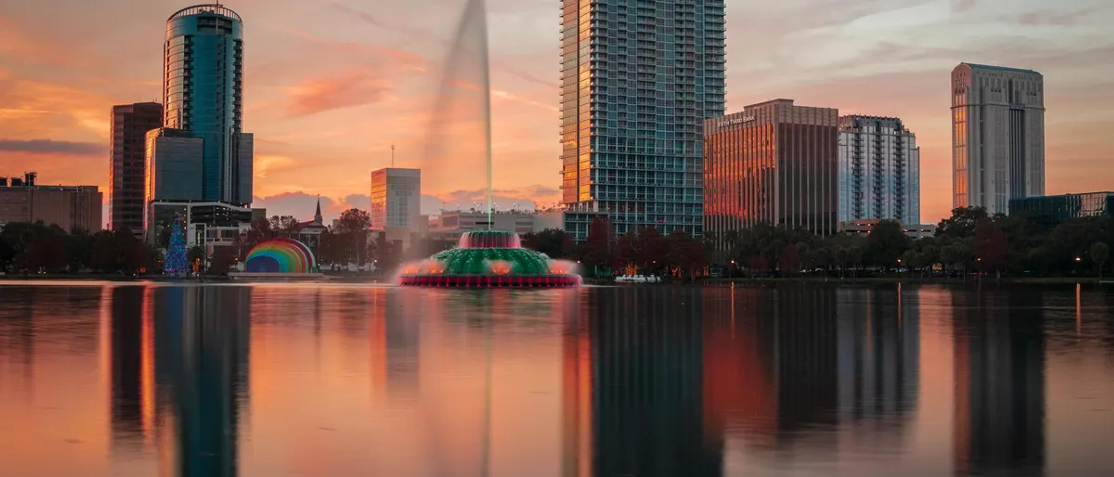 Orlando skyline at sunset over Lake Eola, with the fountain and a rainbow-lit bandshell