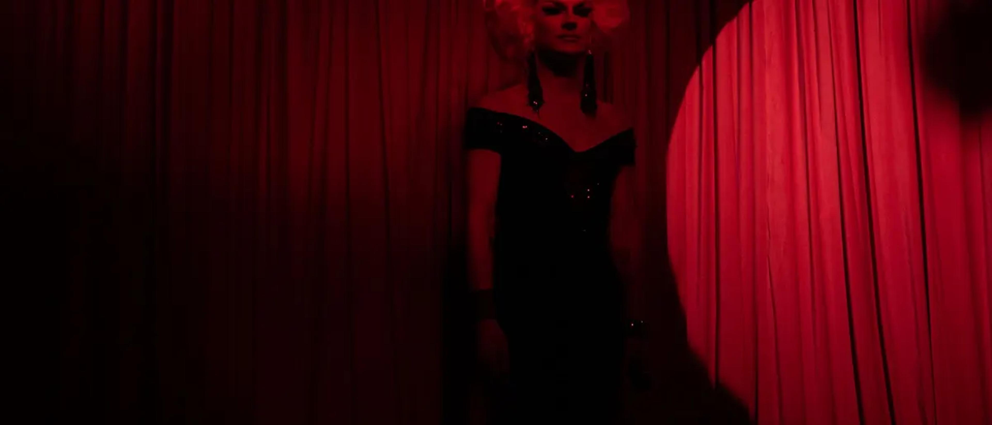 Drag performer in a black gown standing in a red spotlight on a curtained stage