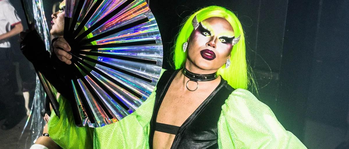 Drag performer in a neon green wig holding a fan under club lights on stage