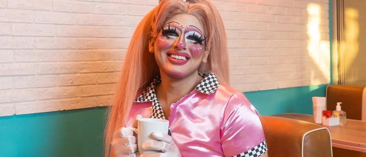 Drag performer in a pink diner outfit smiling with a coffee mug at a bright brunch spot