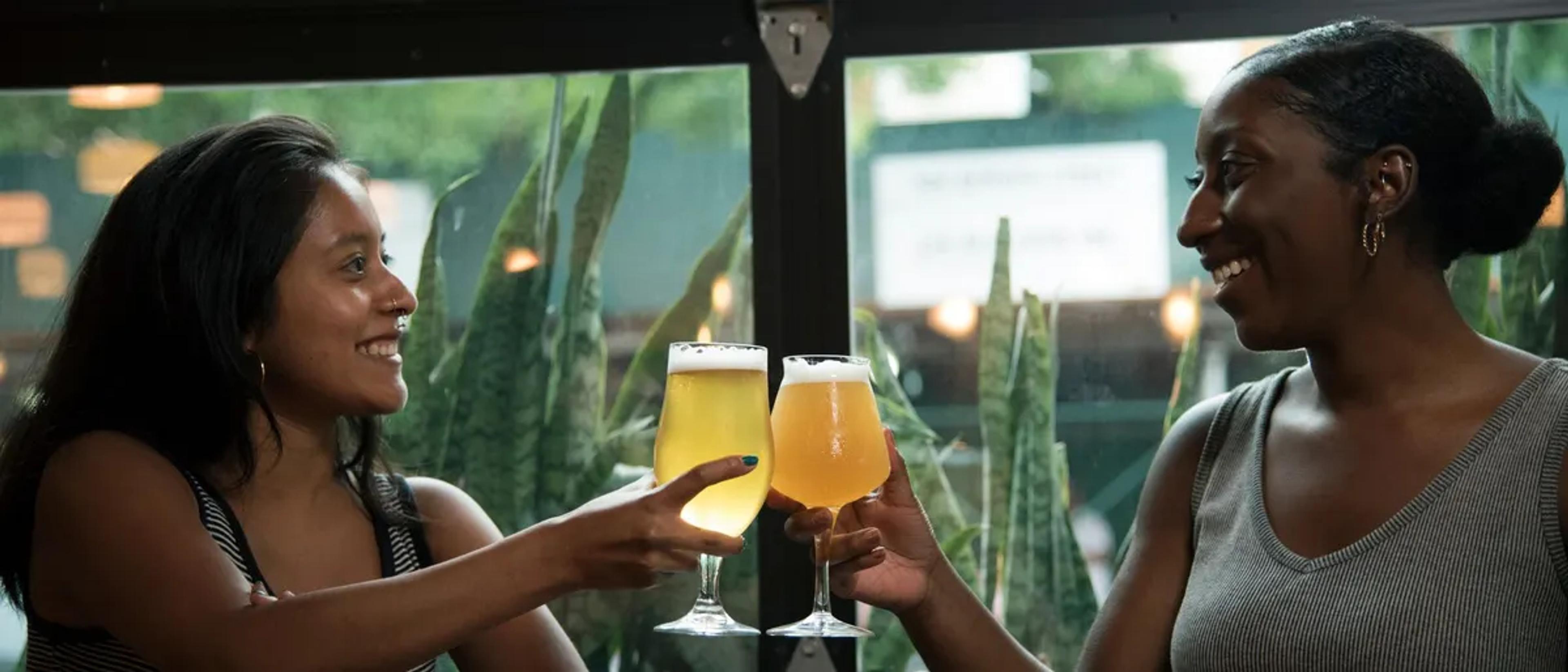 Two women toasting beer glasses by a plant-filled window at a relaxed bar