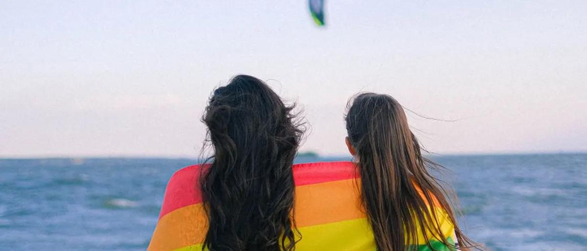 Two people draped in a rainbow flag looking out at the ocean at sunset