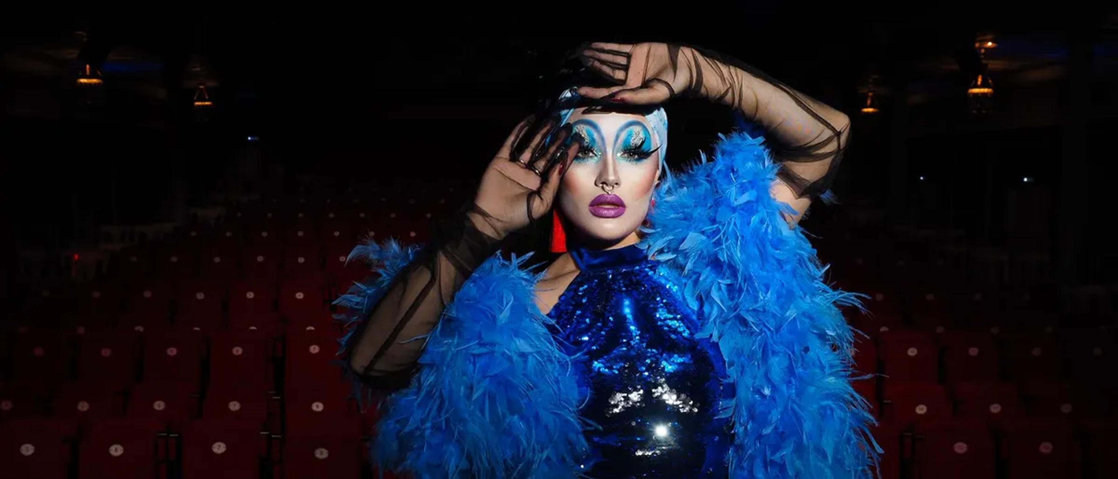 Drag performer in a blue feather boa posing on a theater stage in front of empty red seats