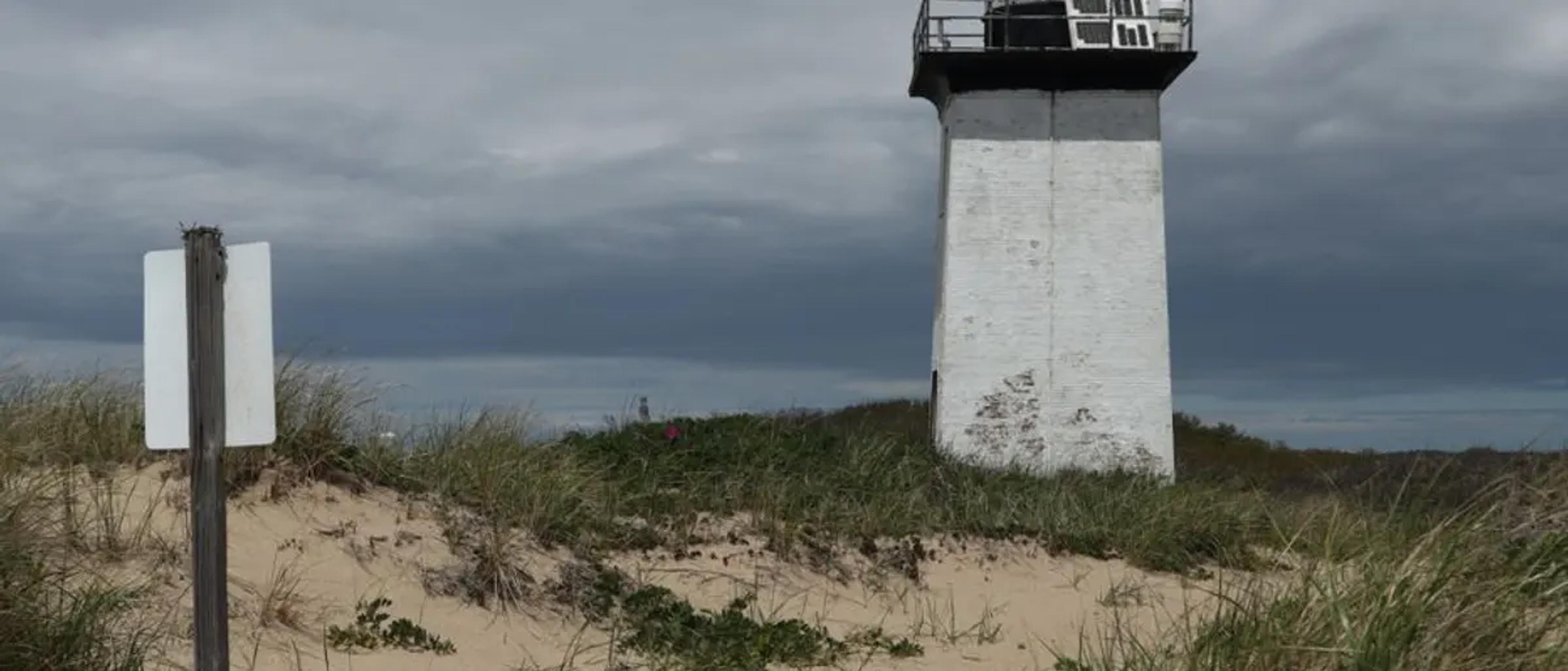 A white lighthouse rising over beach-grass dunes beneath a moody winter sky