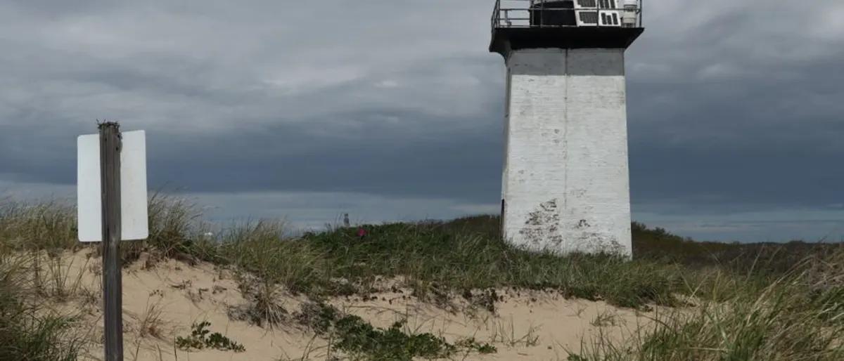 A white lighthouse rising over beach-grass dunes beneath a moody winter sky