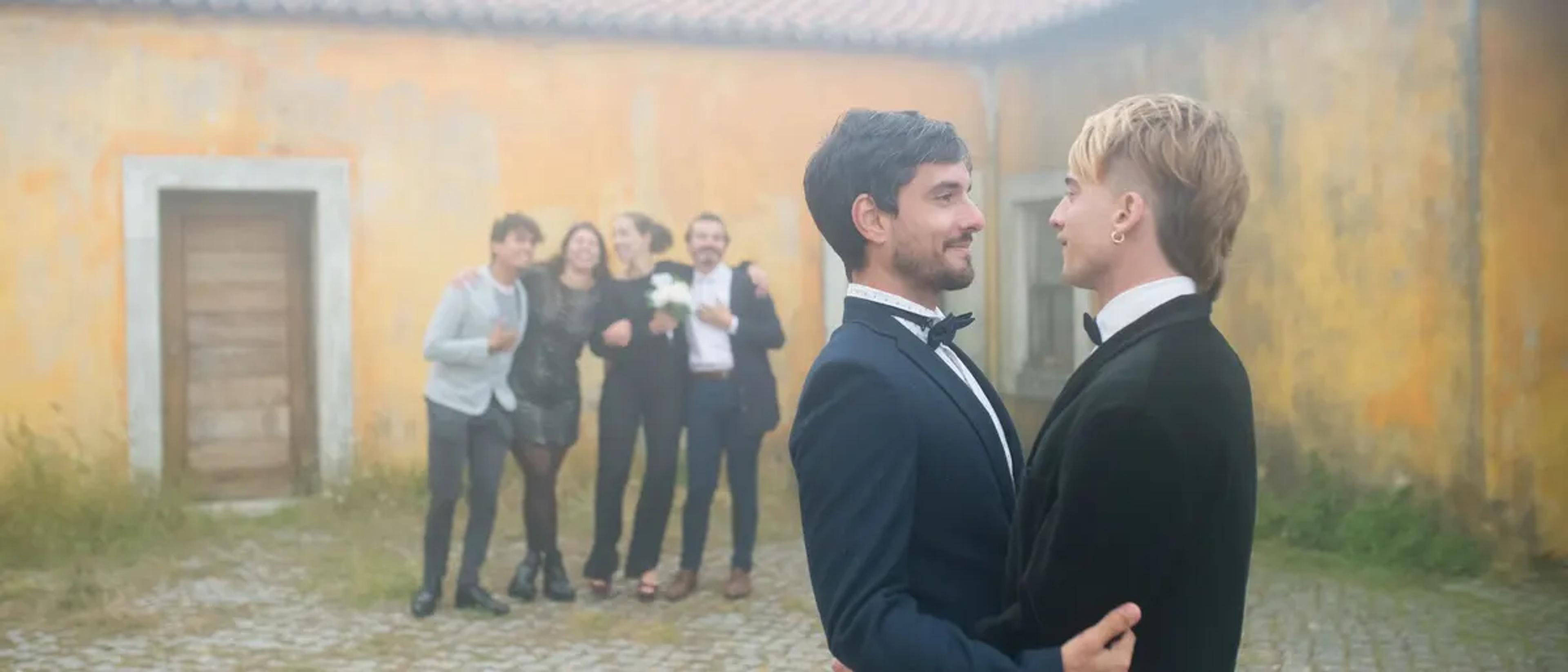 Two grooms in tuxedos embracing at their outdoor wedding with friends behind them