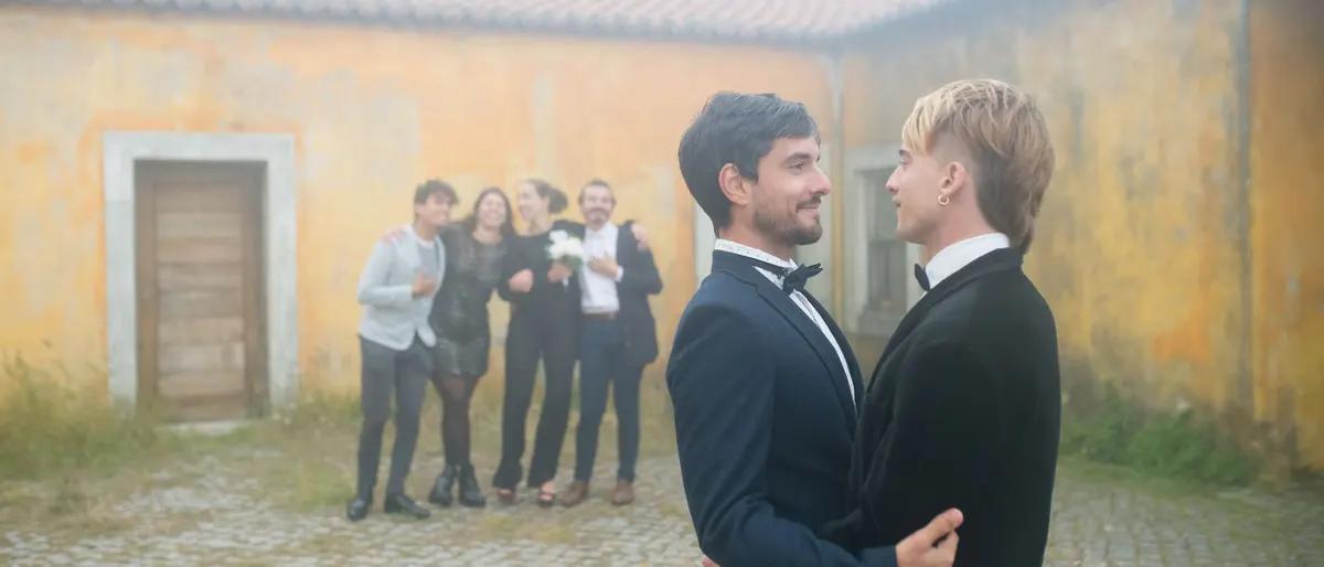 Two grooms in tuxedos embracing at their outdoor wedding with friends behind them