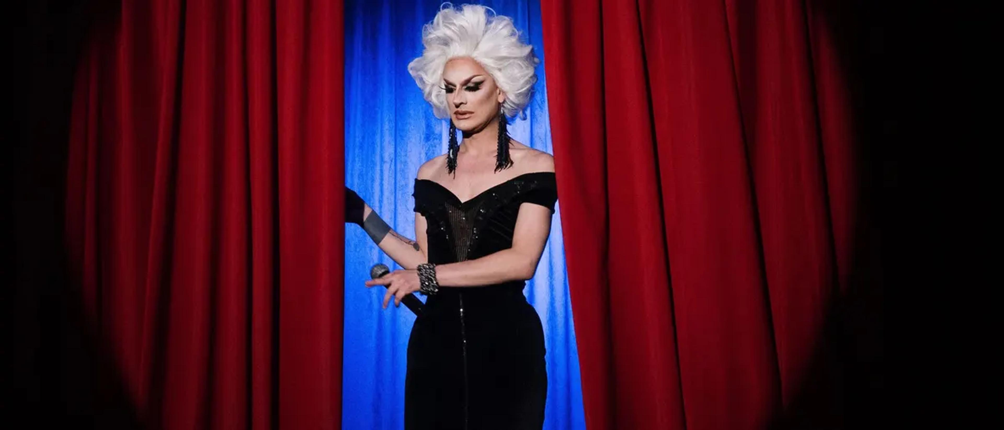 Drag performer in a white wig and black gown stepping through red stage curtains under a spotlight