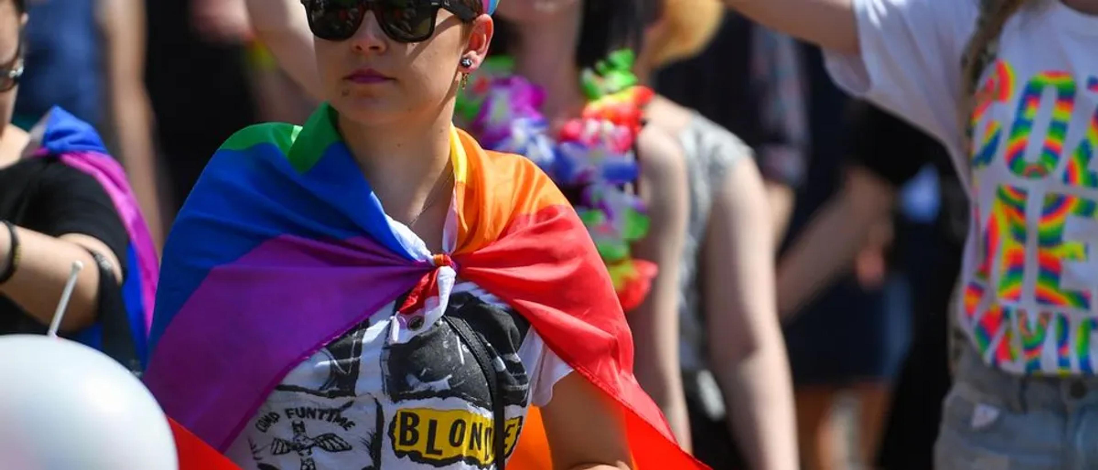 A person draped in a rainbow flag and sunglasses smiling amid a sunny Pride crowd