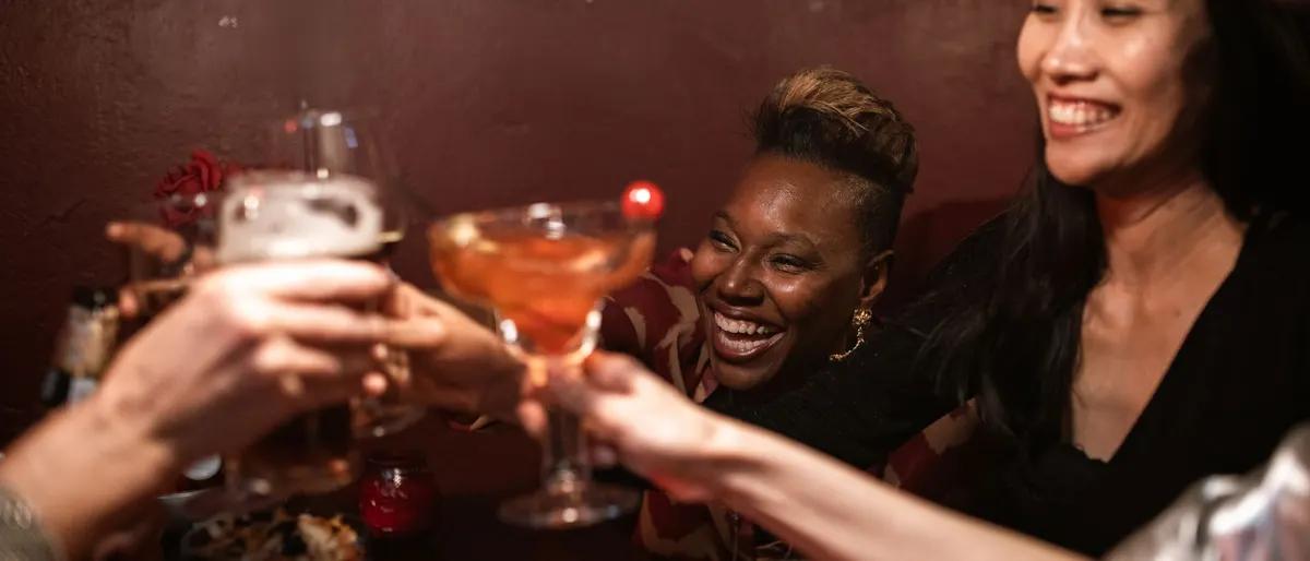 Two women laughing and clinking cocktail glasses together at a warm neighborhood bar