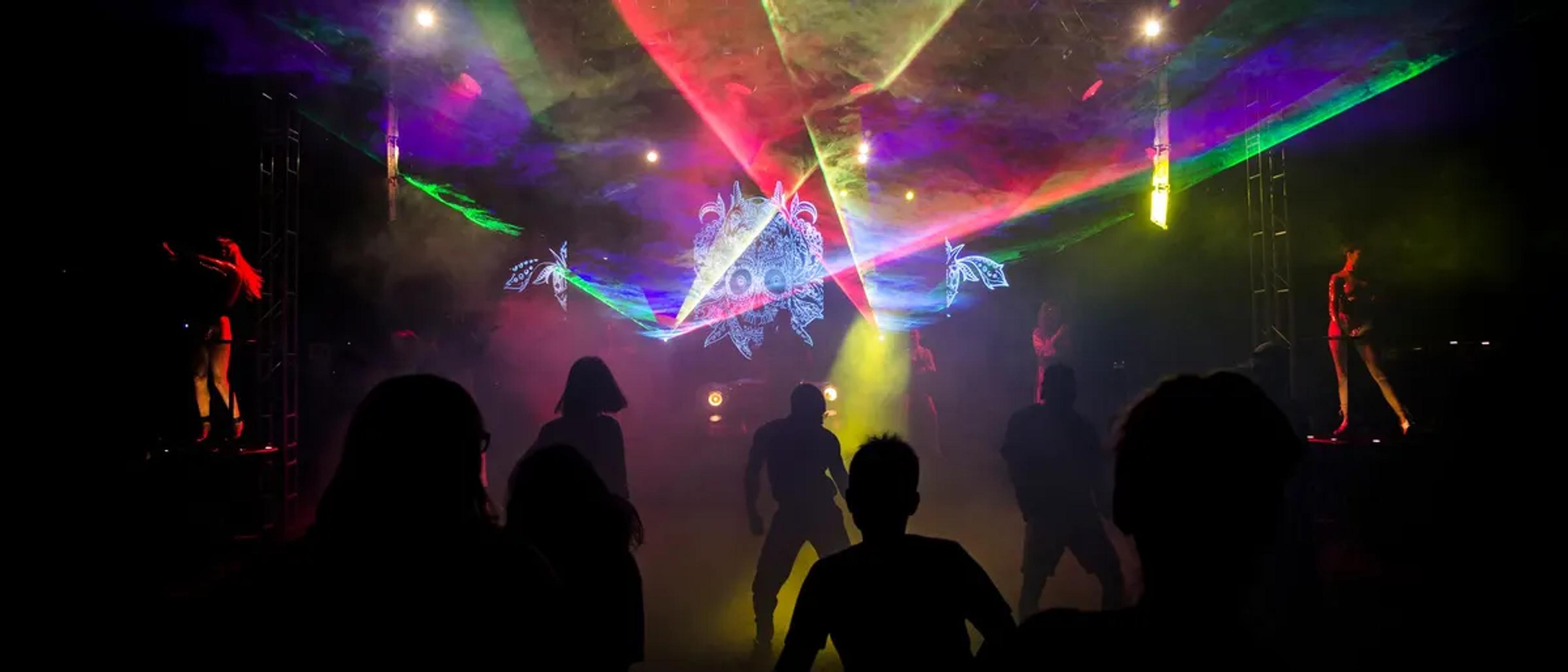 Crowd silhouetted against colorful rainbow stage lasers at a Miami circuit party