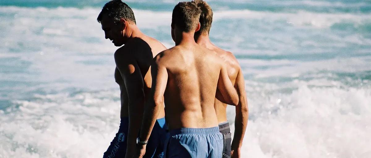 Shirtless men standing together at the ocean's edge looking out at the surf