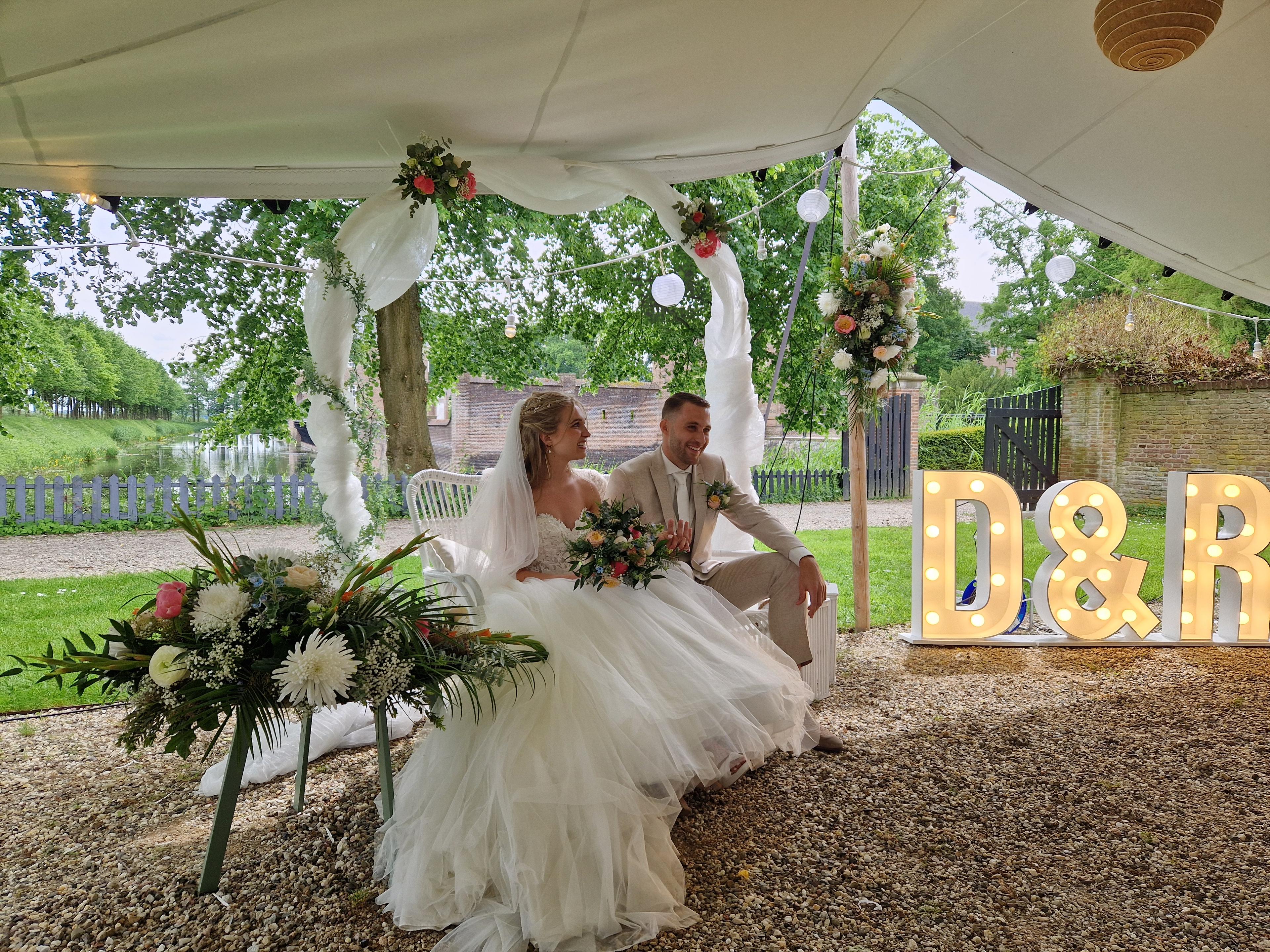 Bruidspaar zit tijdens de ceremonie onder een tentdoek, naast lichtletters D&R