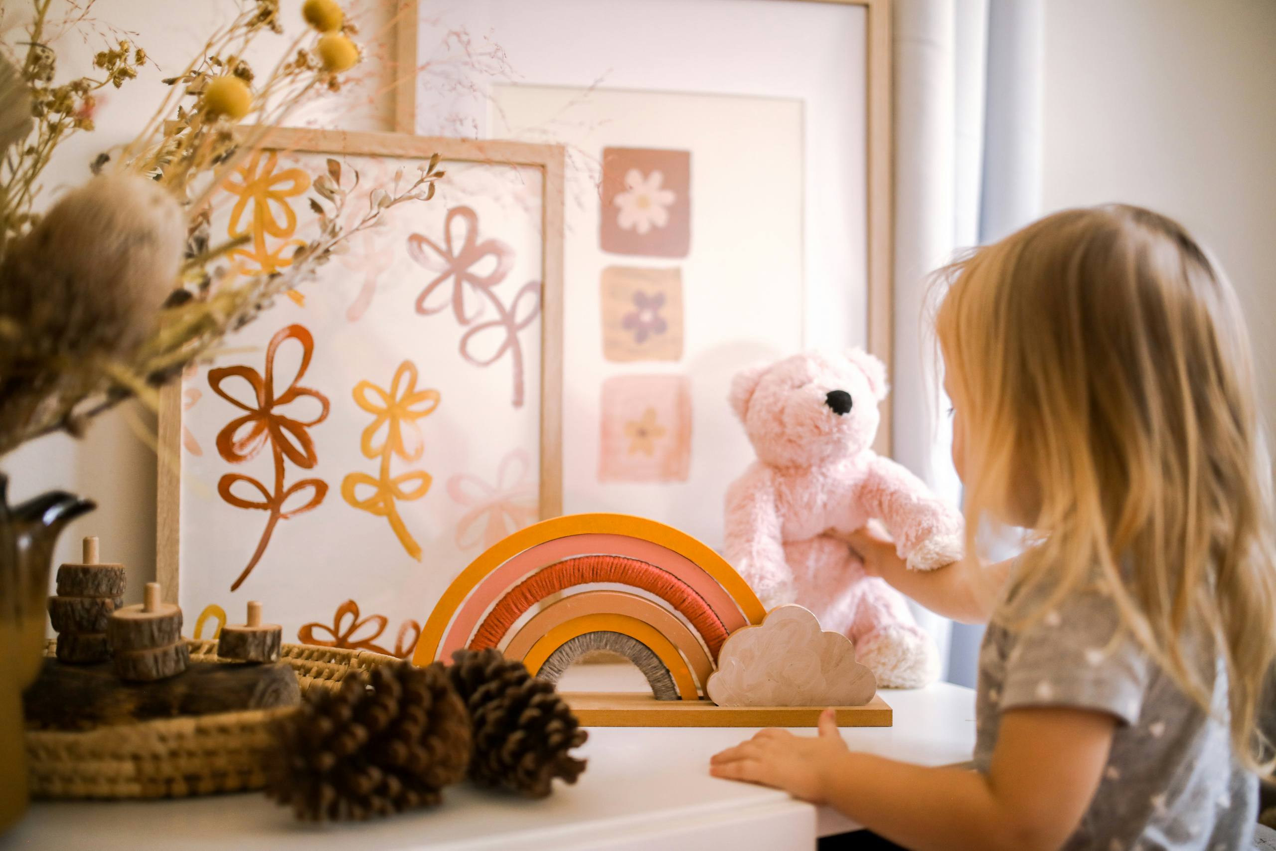 A child playing beside a shelf of soft toys, a wooden rainbow and framed drawings