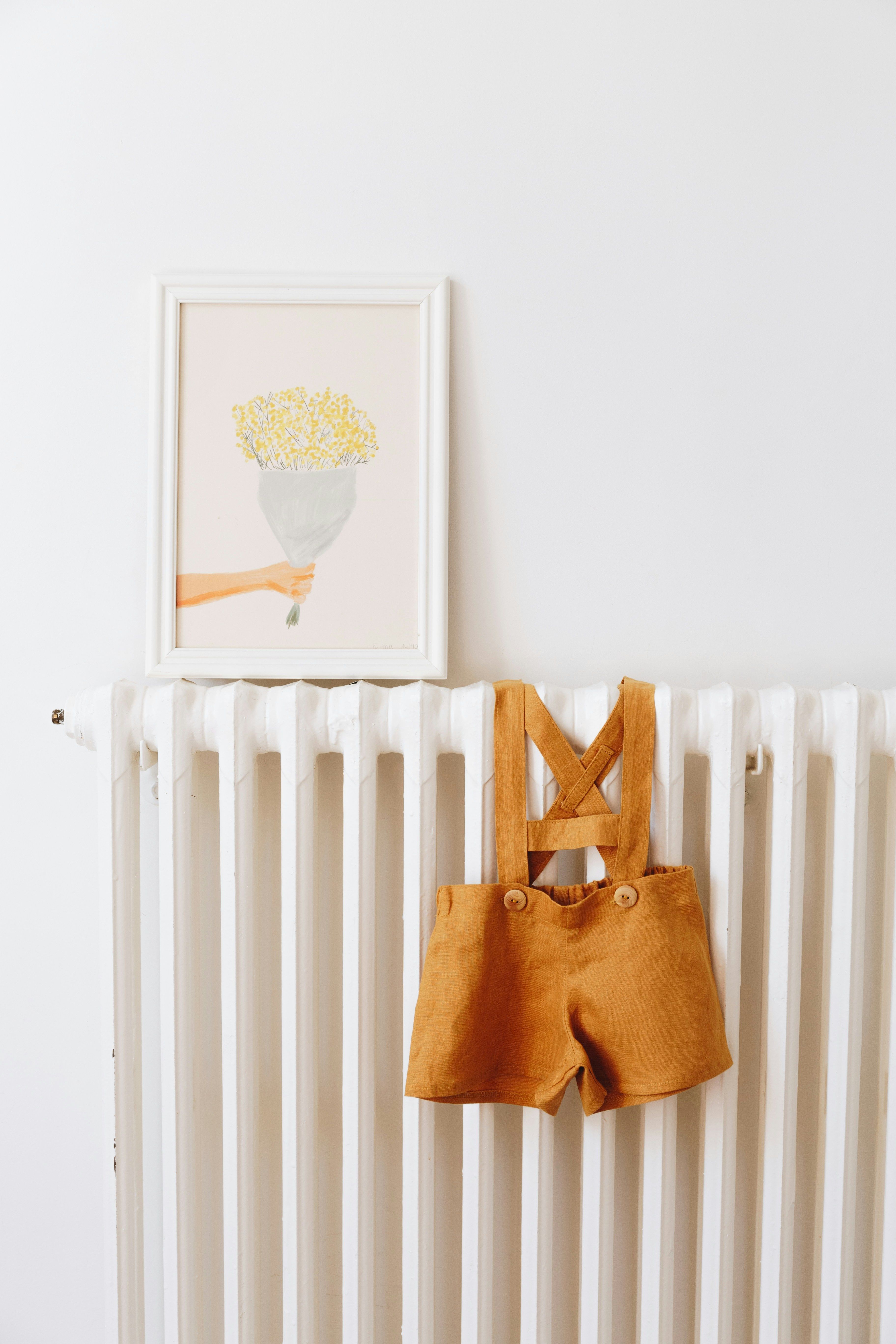 A child's mustard dungarees hanging on a radiator below a framed print